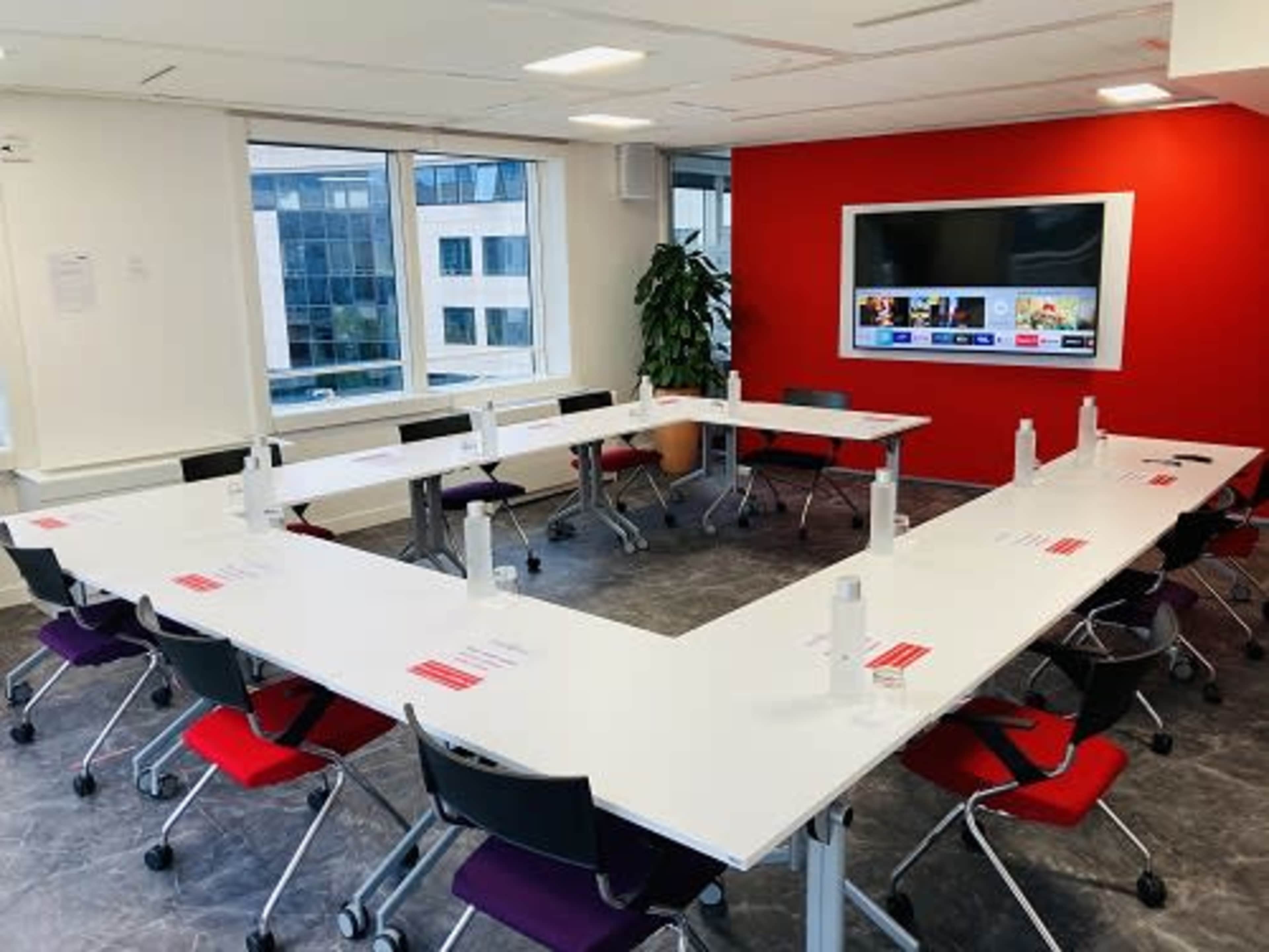 A modern conference room features a U-shaped arrangement of white tables and colorful chairs, with a large screen mounted on a red accent wall.