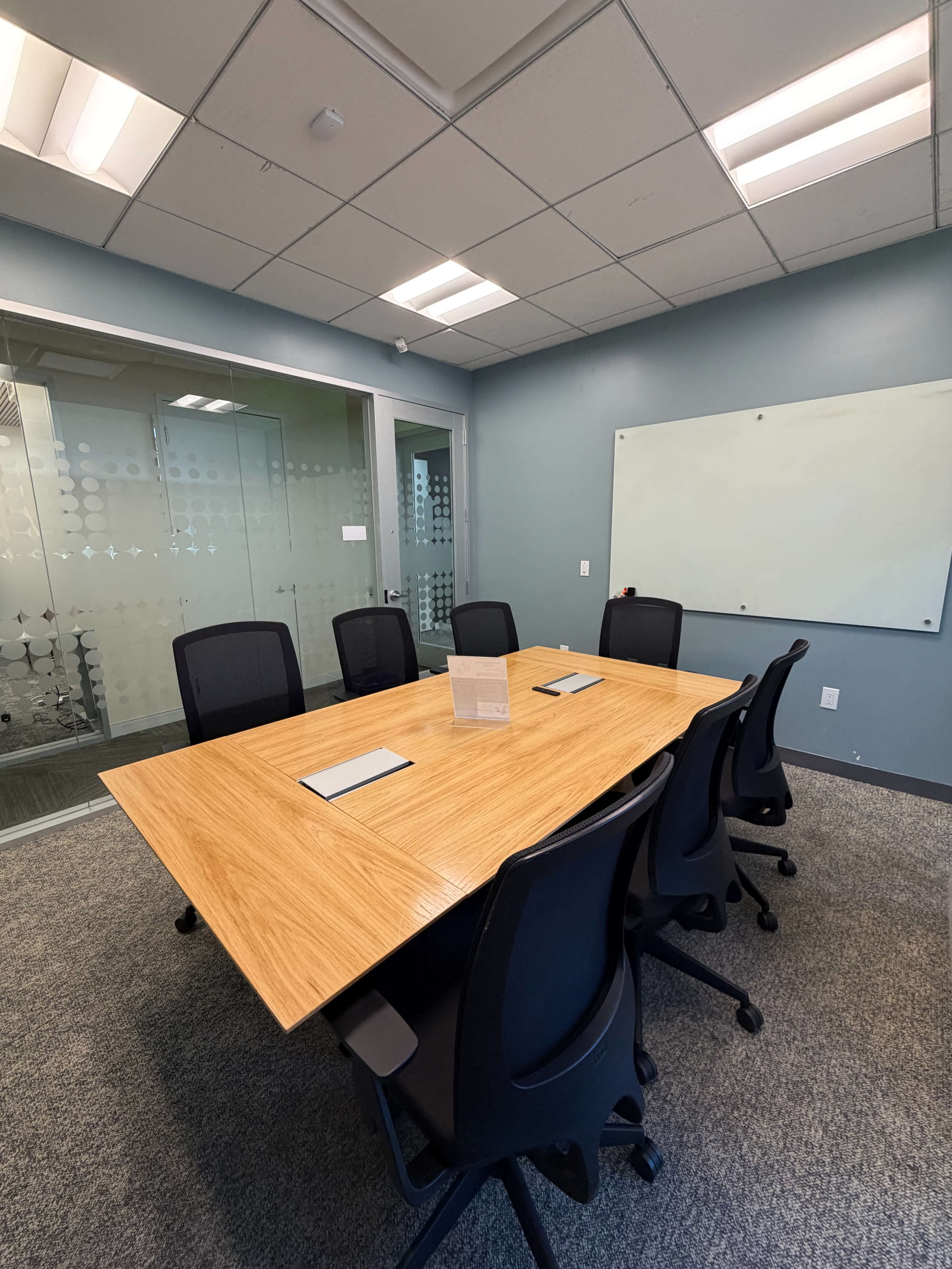 The image shows a conference room with a large wooden table surrounded by black chairs, a glass door, and a whiteboard on the wall.
