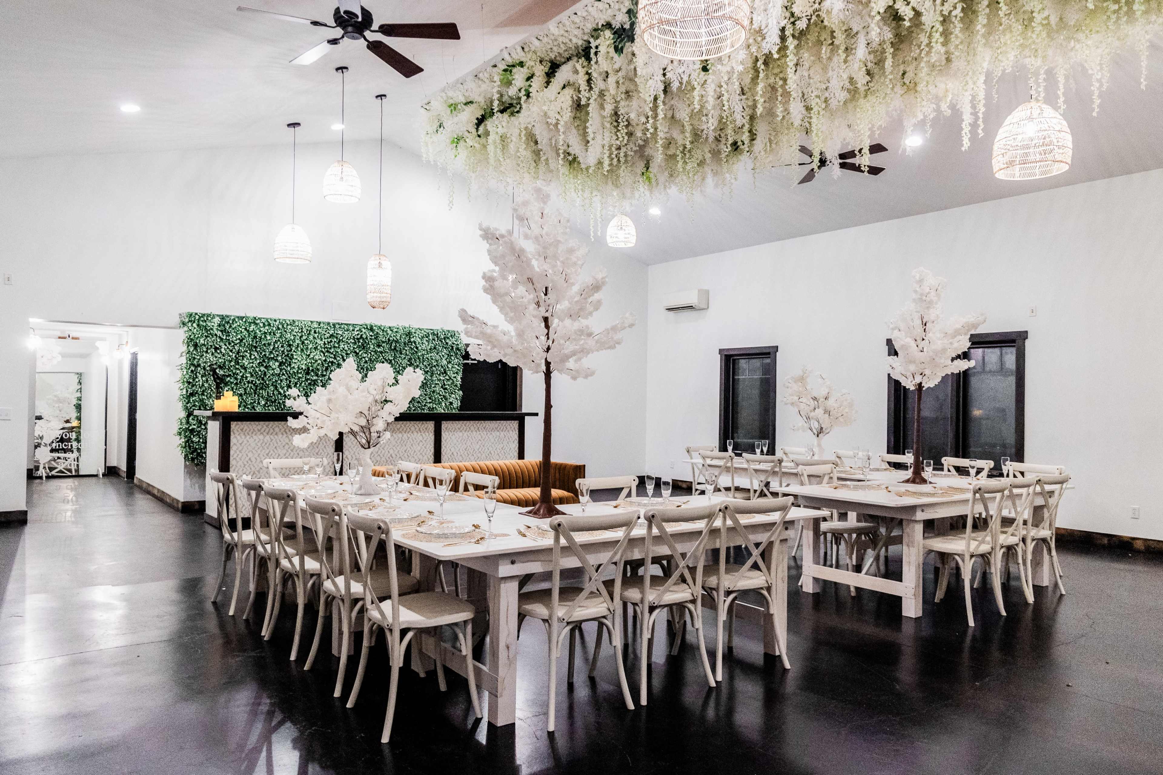 A spacious dining area features long, white tables surrounded by chairs, with faux trees and floral decorations overhead.