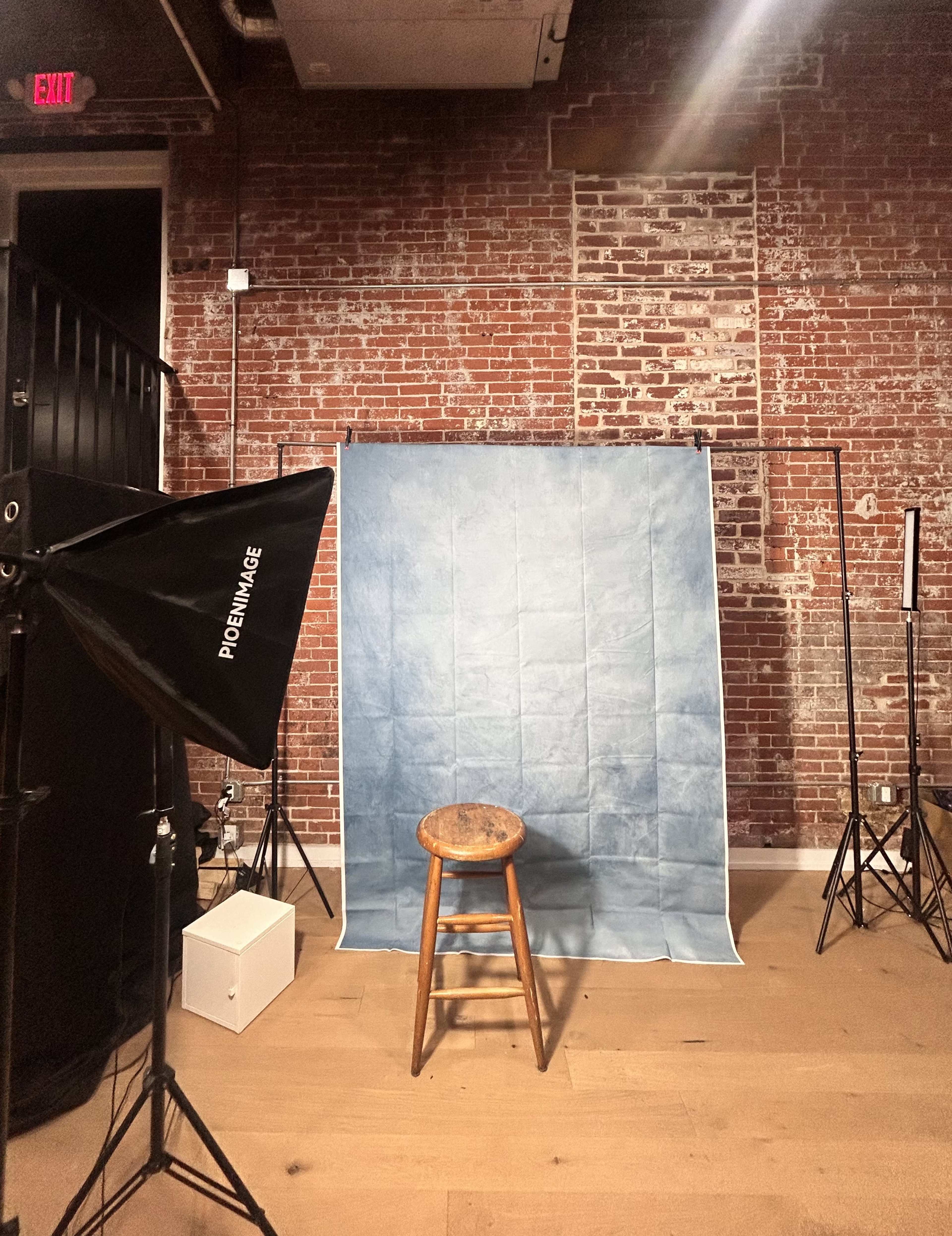 The image shows a photography studio setup with a blue backdrop, a wooden stool in front, and studio lighting equipment on either side.