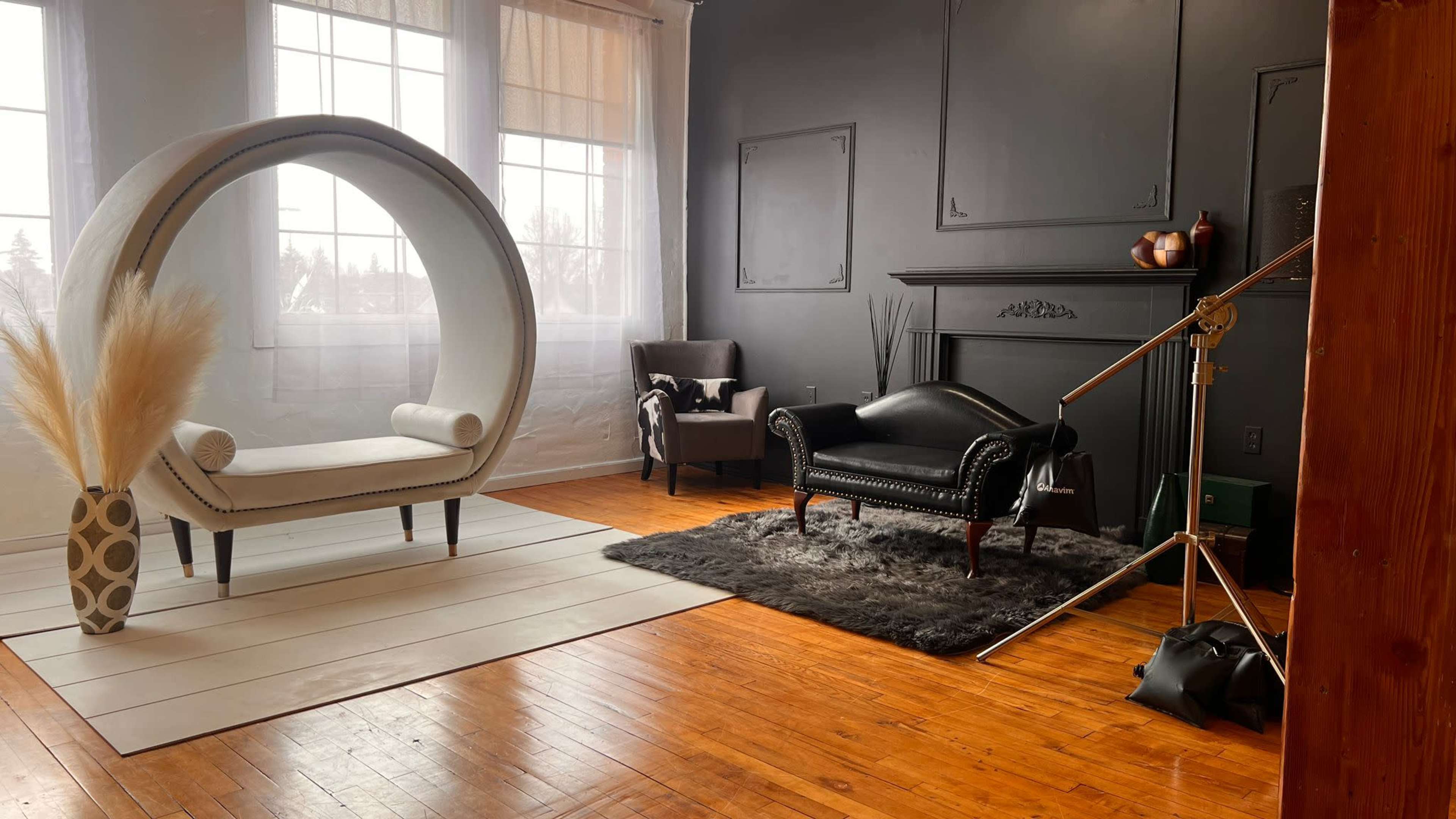 The room features a circular white chair on a large area rug, a black leather sofa, and various decorative elements against dark walls and large windows.