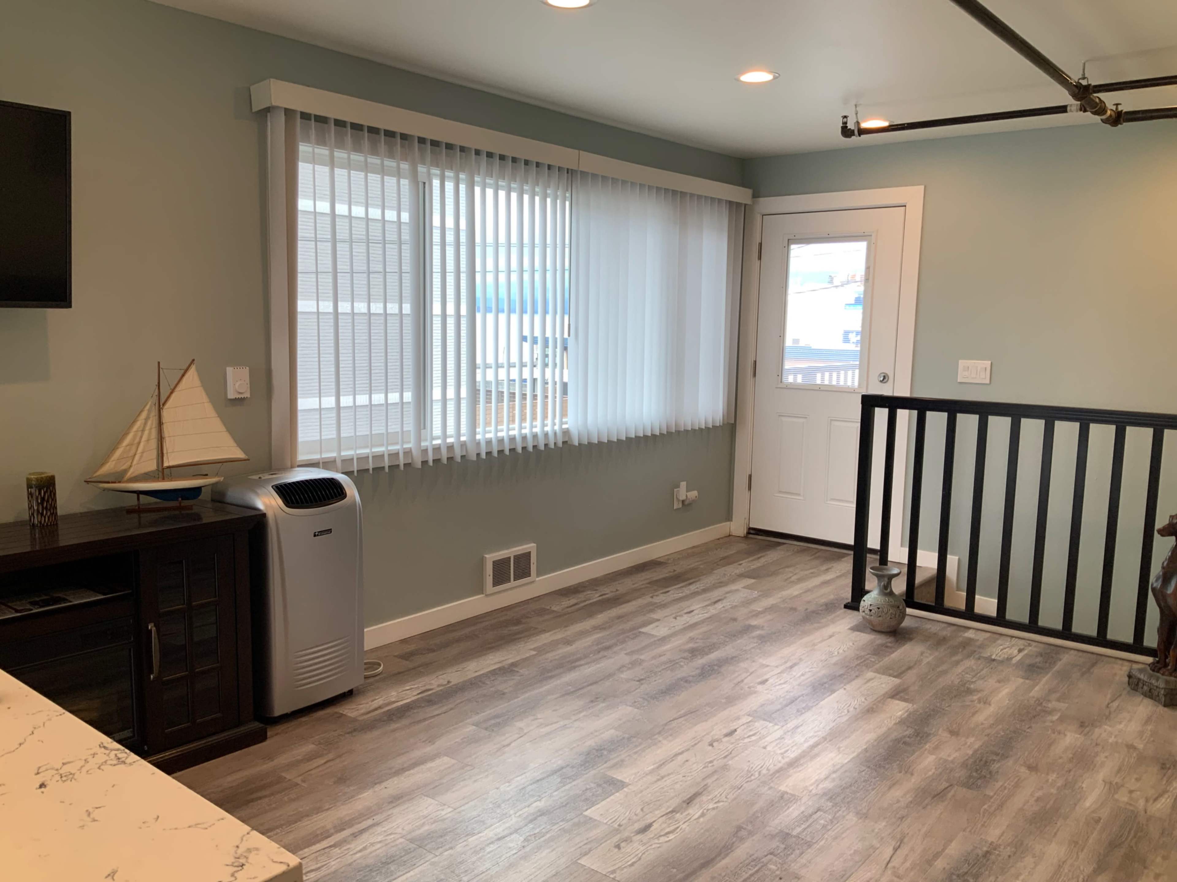 A room with light-colored walls, wooden flooring, a window with vertical blinds, and a door leading outside.