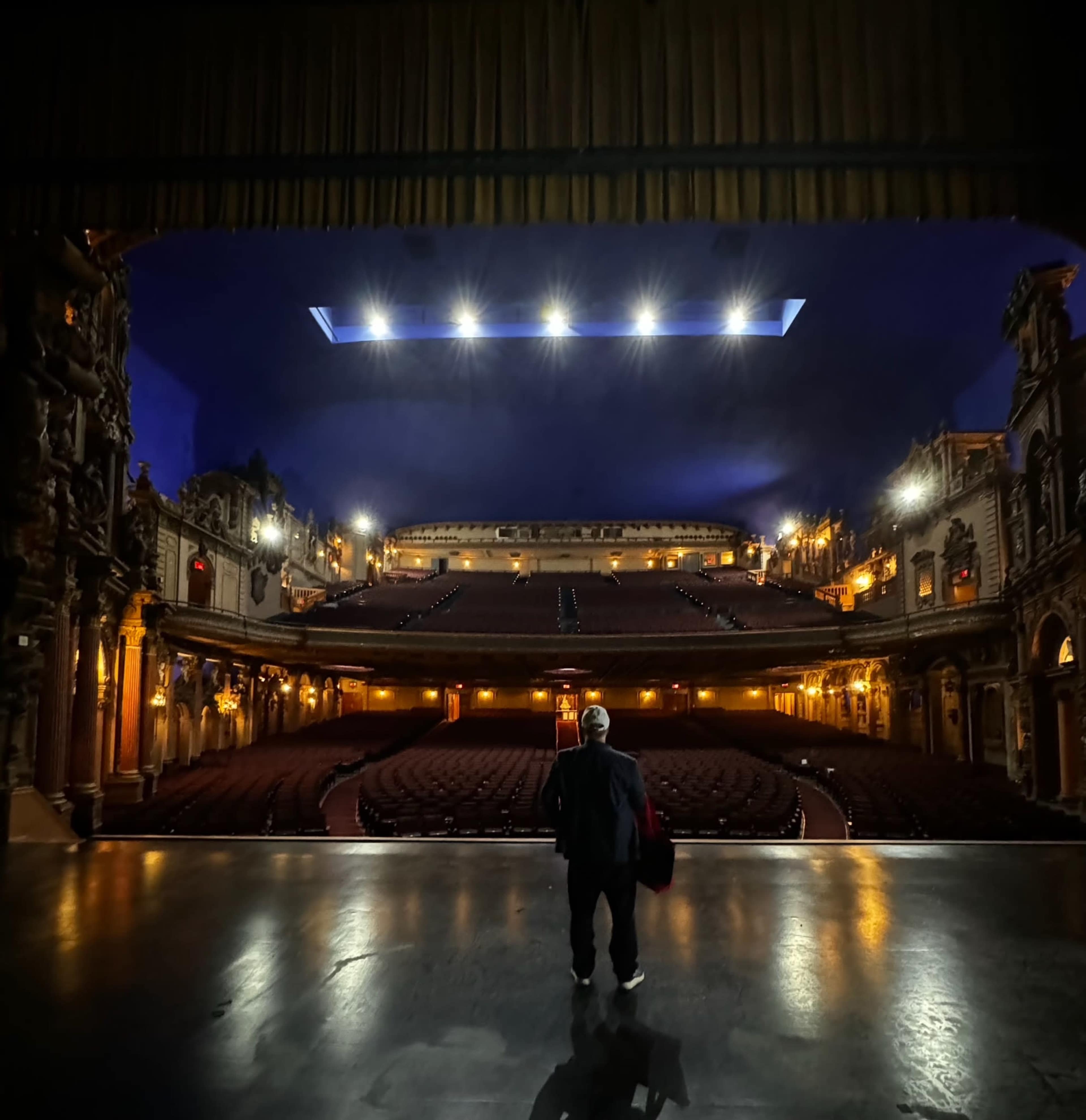 A person stands on stage looking out at an empty theater with ornate architecture and dim lighting.