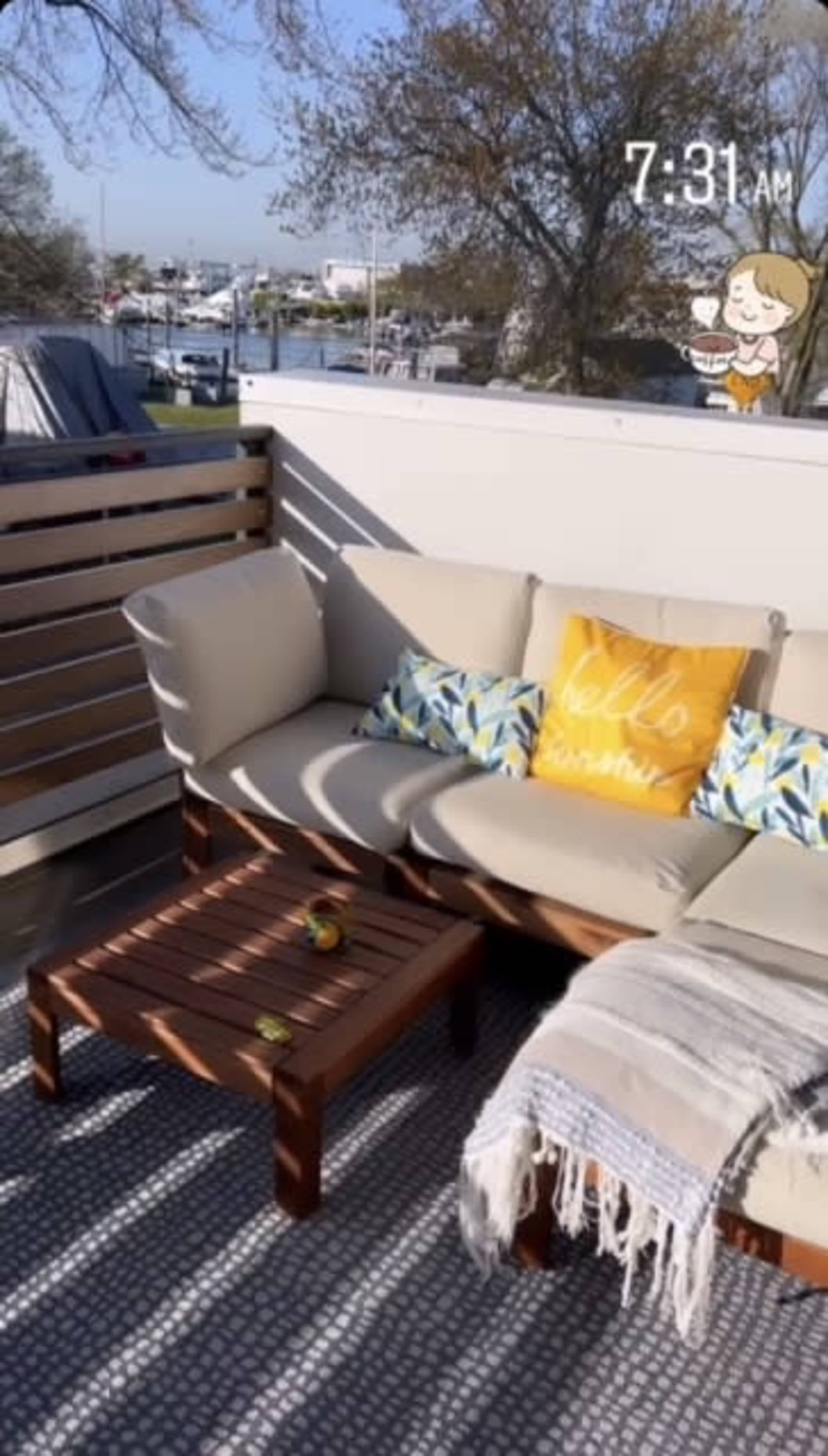 The image shows a cozy balcony with a beige couch adorned with decorative pillows, a wooden coffee table, and a soft blanket, set against a bright blue sky.