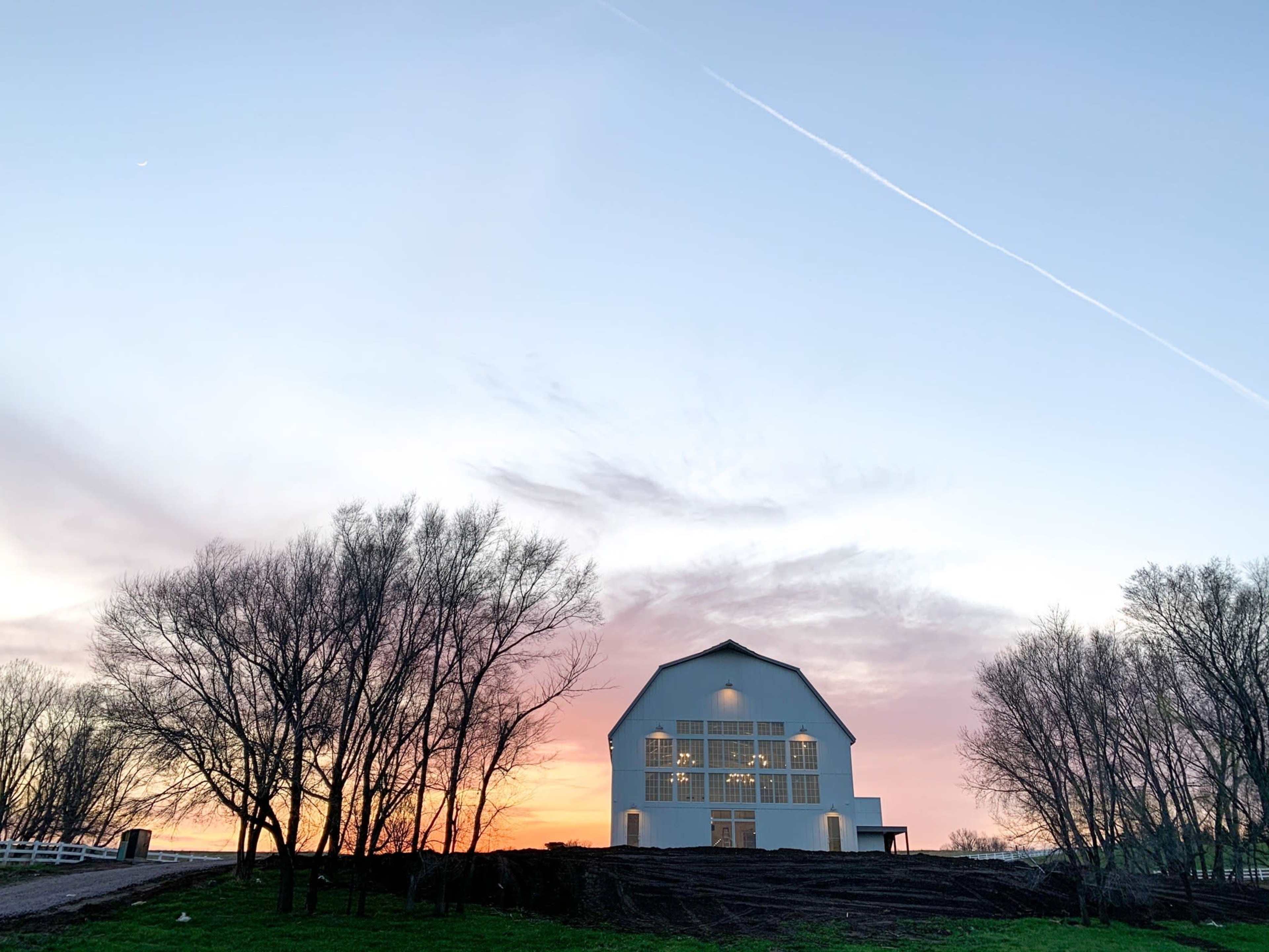 A white barn stands against a colorful sunset sky, framed by bare trees.