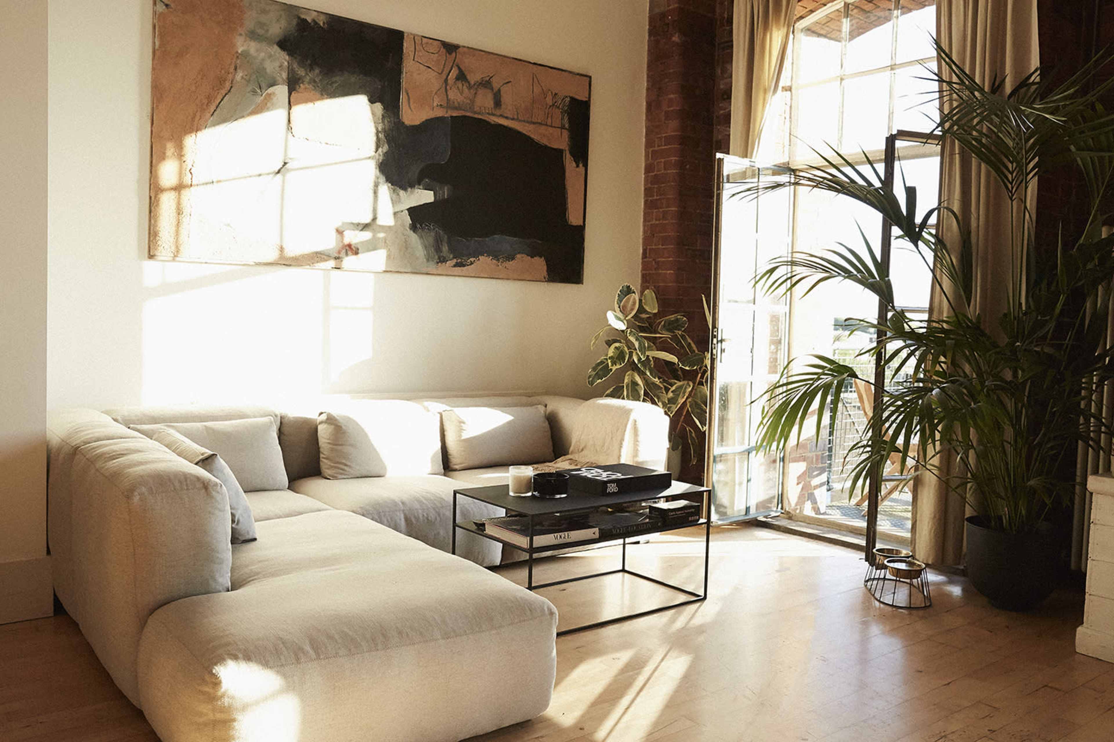 A modern living room features a large, beige sectional sofa, a glass-top coffee table, and a potted plant, with sunlight streaming in through expansive windows.