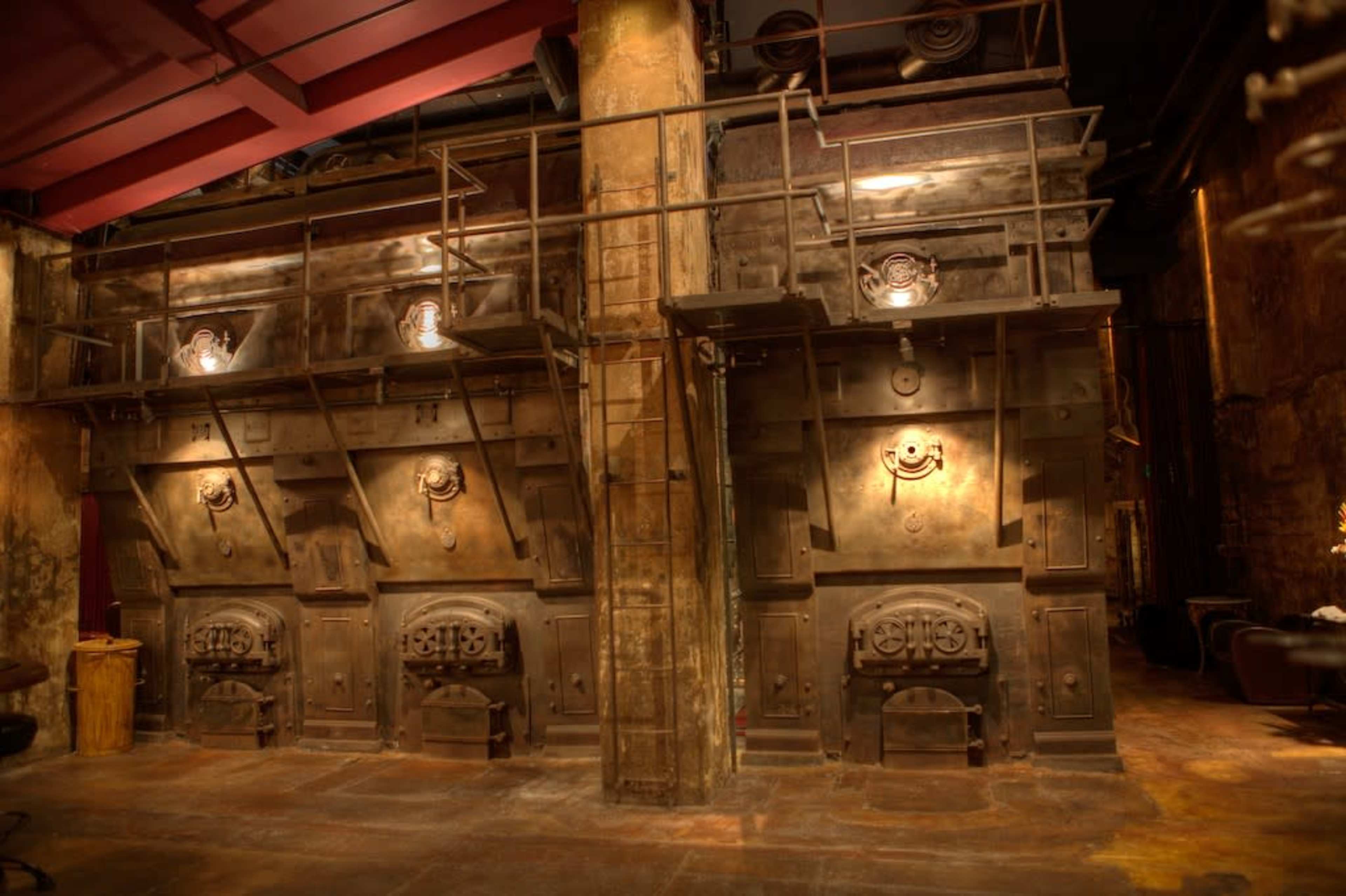 The image shows a dimly lit industrial space with large metal ovens mounted on a wall, featuring intricate designs and overhead lighting.