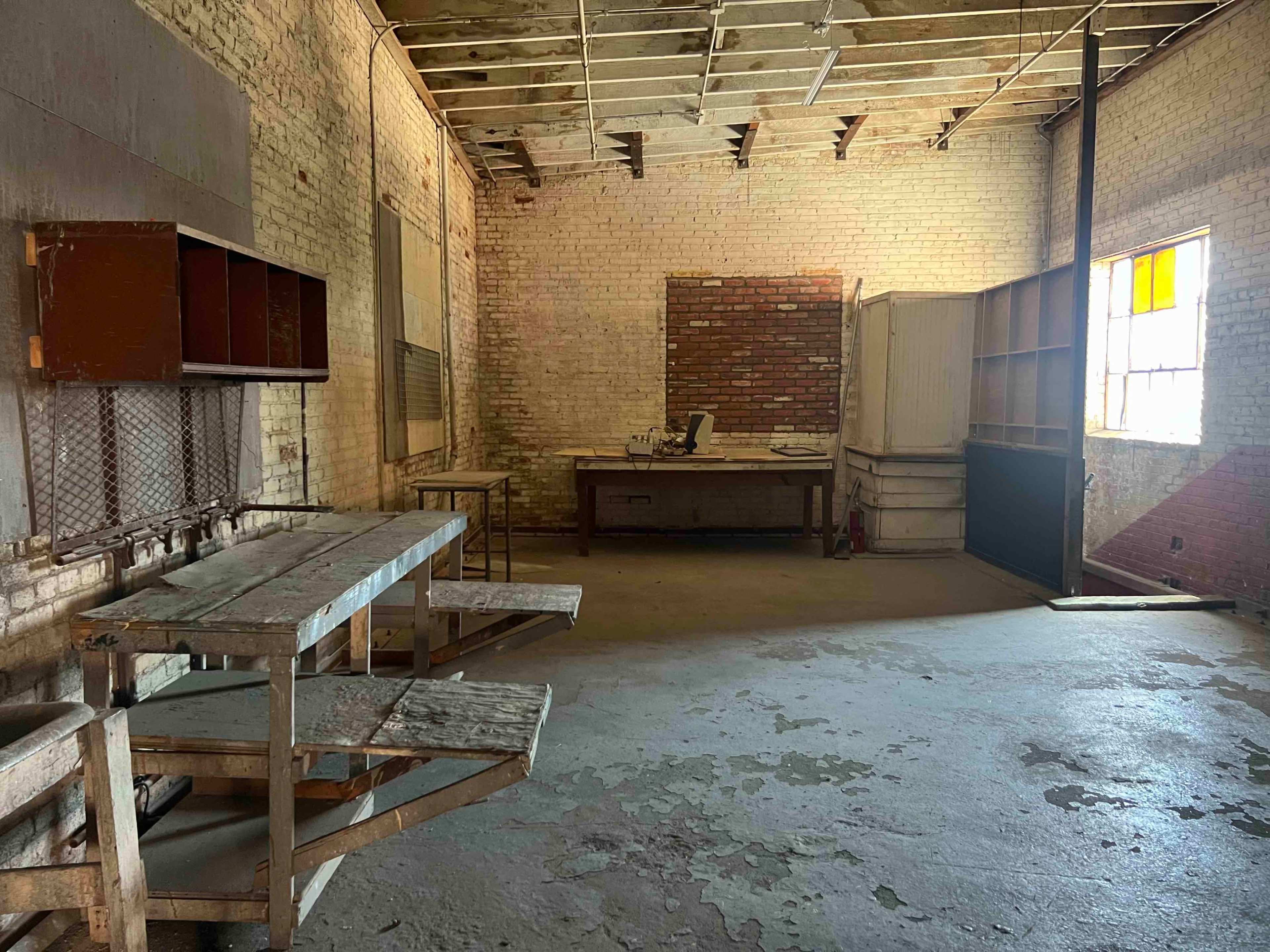 An empty, bare room with exposed brick walls, featuring a wooden table, a workbench, and a window allowing light to enter.