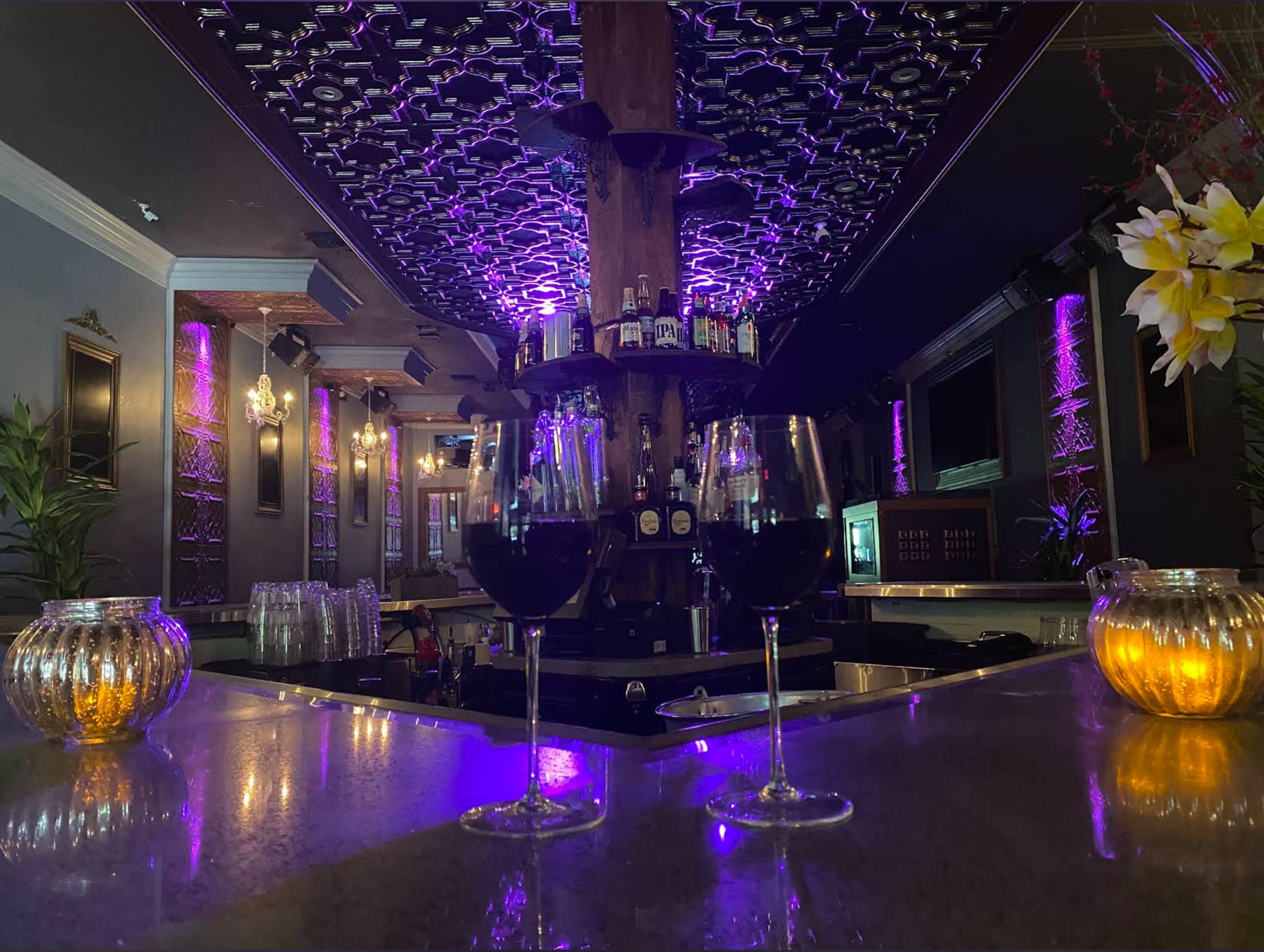A dimly lit bar features two glasses of red wine on a polished countertop, with shelves of bottles and decorative lights in the background.