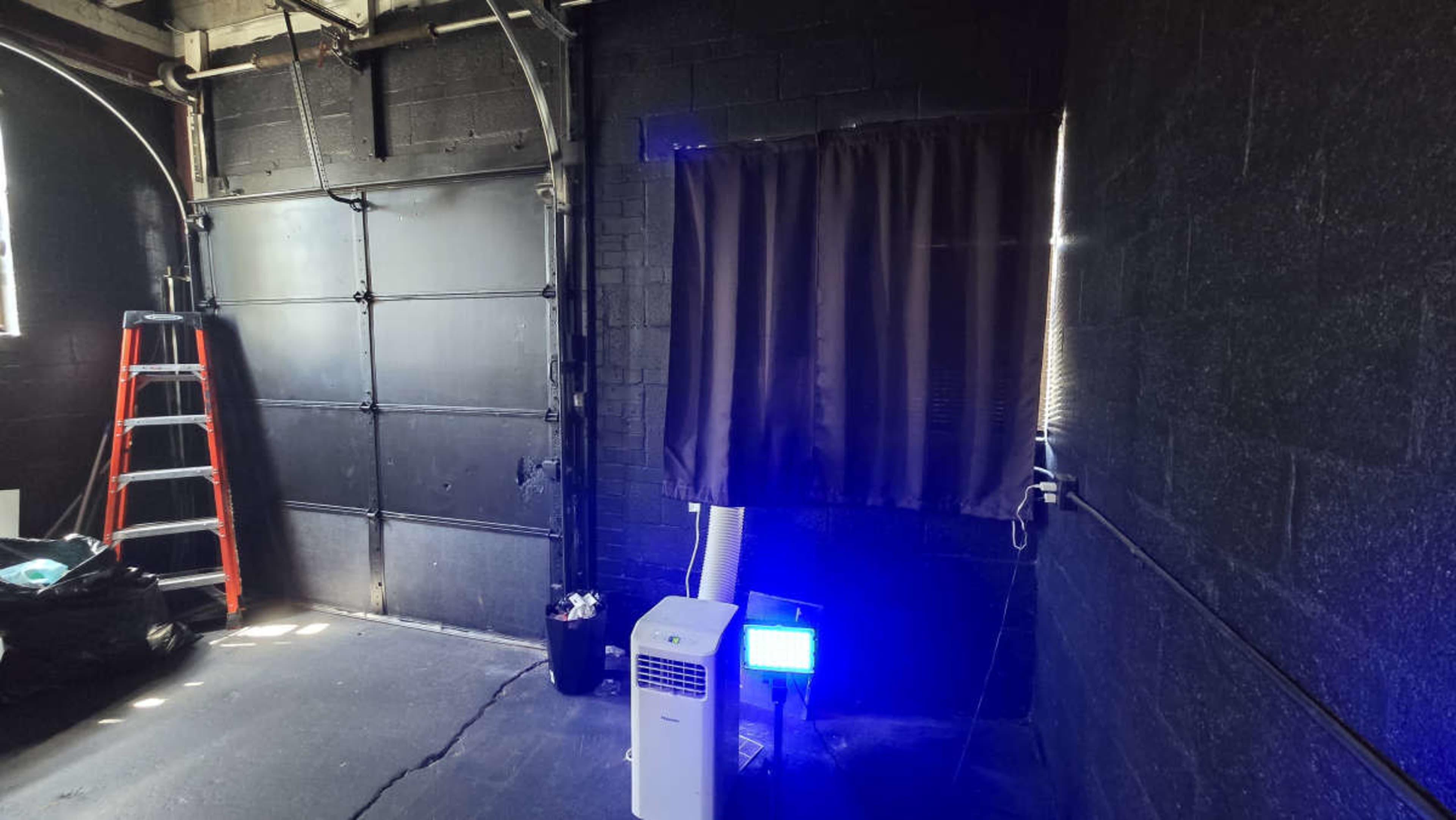 The image shows a dimly lit garage with a black wall, a closed garage door, a ladder leaning against the wall, a portable air conditioner, and a blue light near a curtained window.