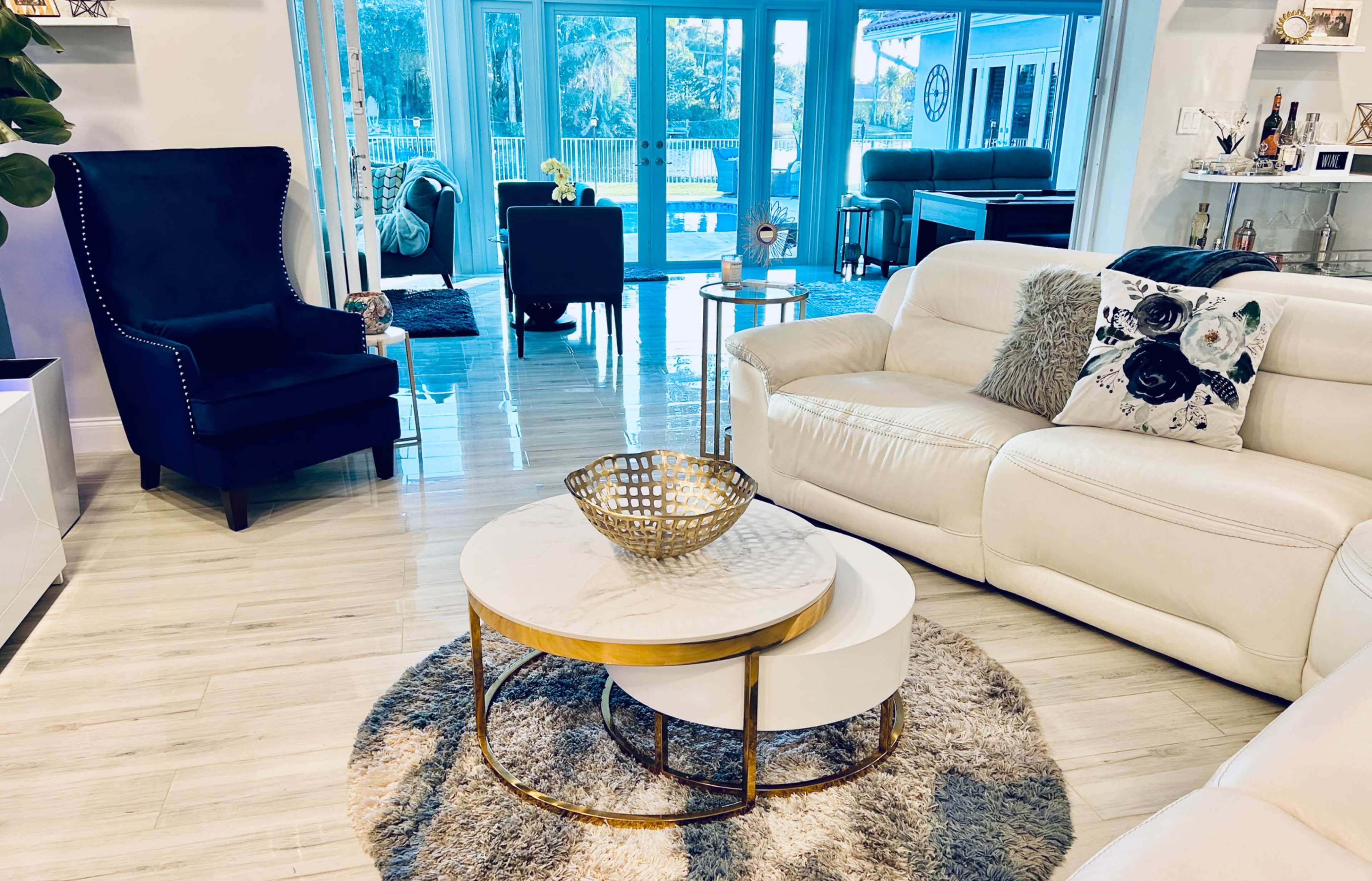 The living room features a white leather sofa, a round coffee table, a plush rug, and large windows overlooking a patio with outdoor furniture.