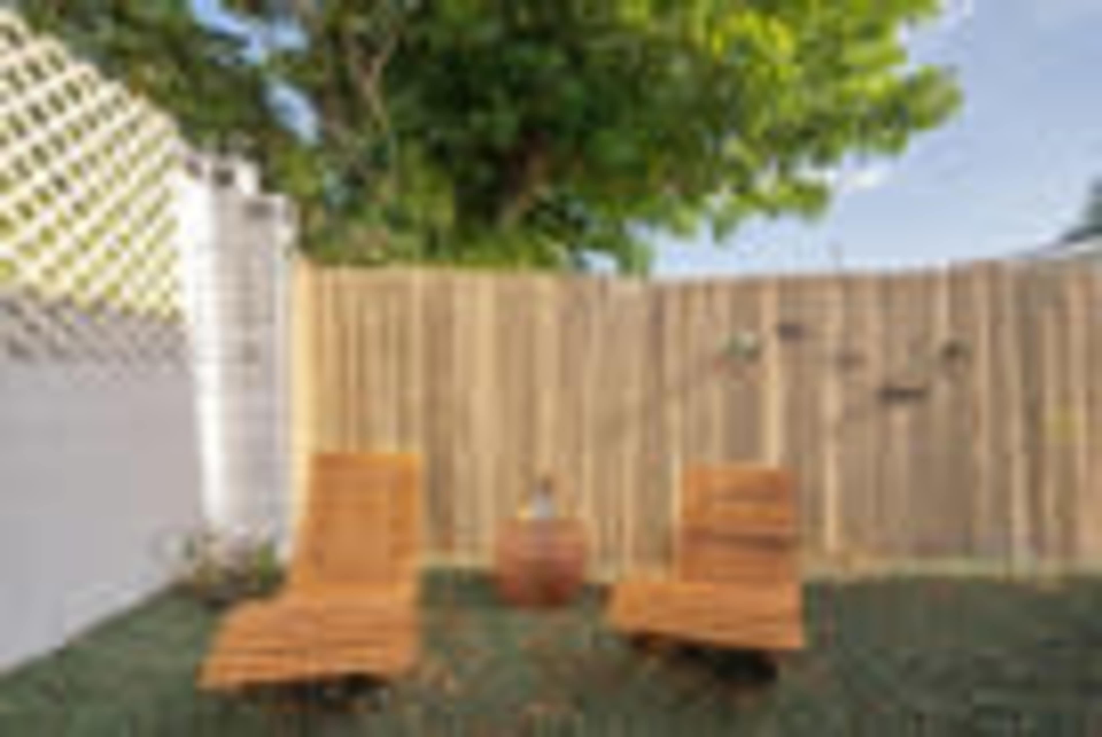 Two wooden lounge chairs and a small round table are positioned on a grassy area in a private outdoor space with a wooden fence and a tree overhead.