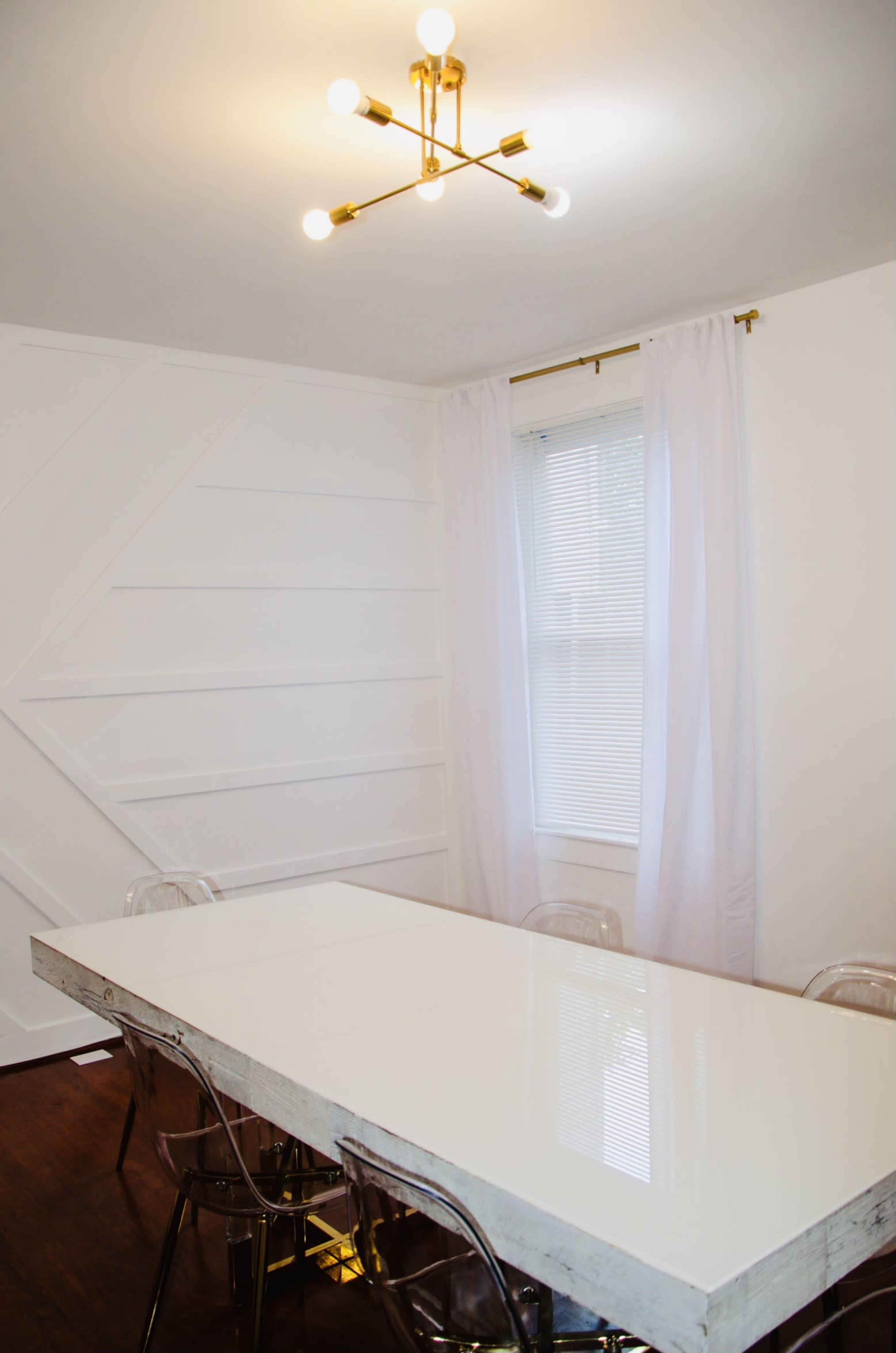 A modern dining room features a white table surrounded by clear chairs under a geometric ceiling light and a window with sheer curtains.