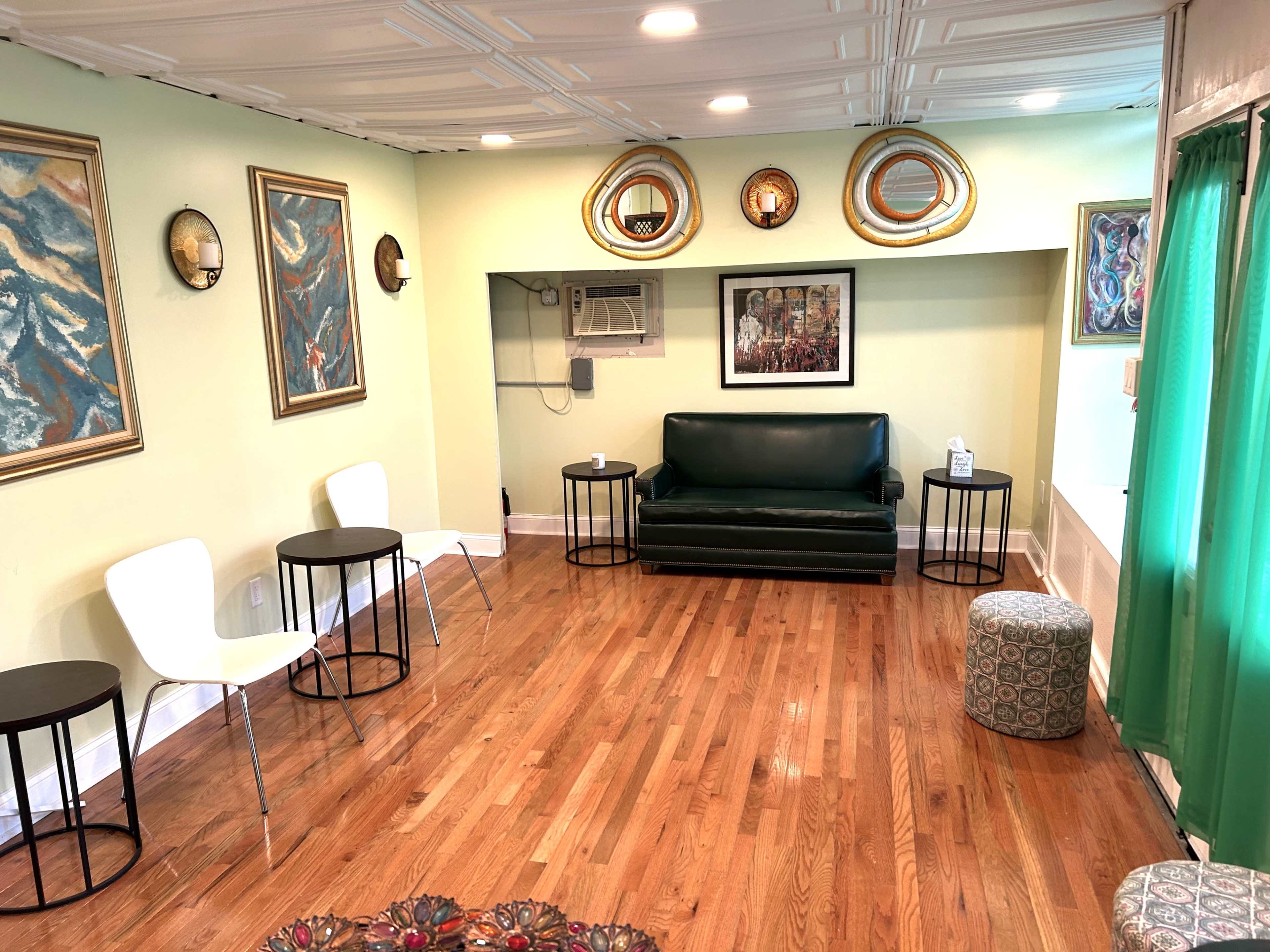 The room features a green sofa, two white chairs, small black tables, and several framed artworks on the walls, with wooden flooring and a green curtain.