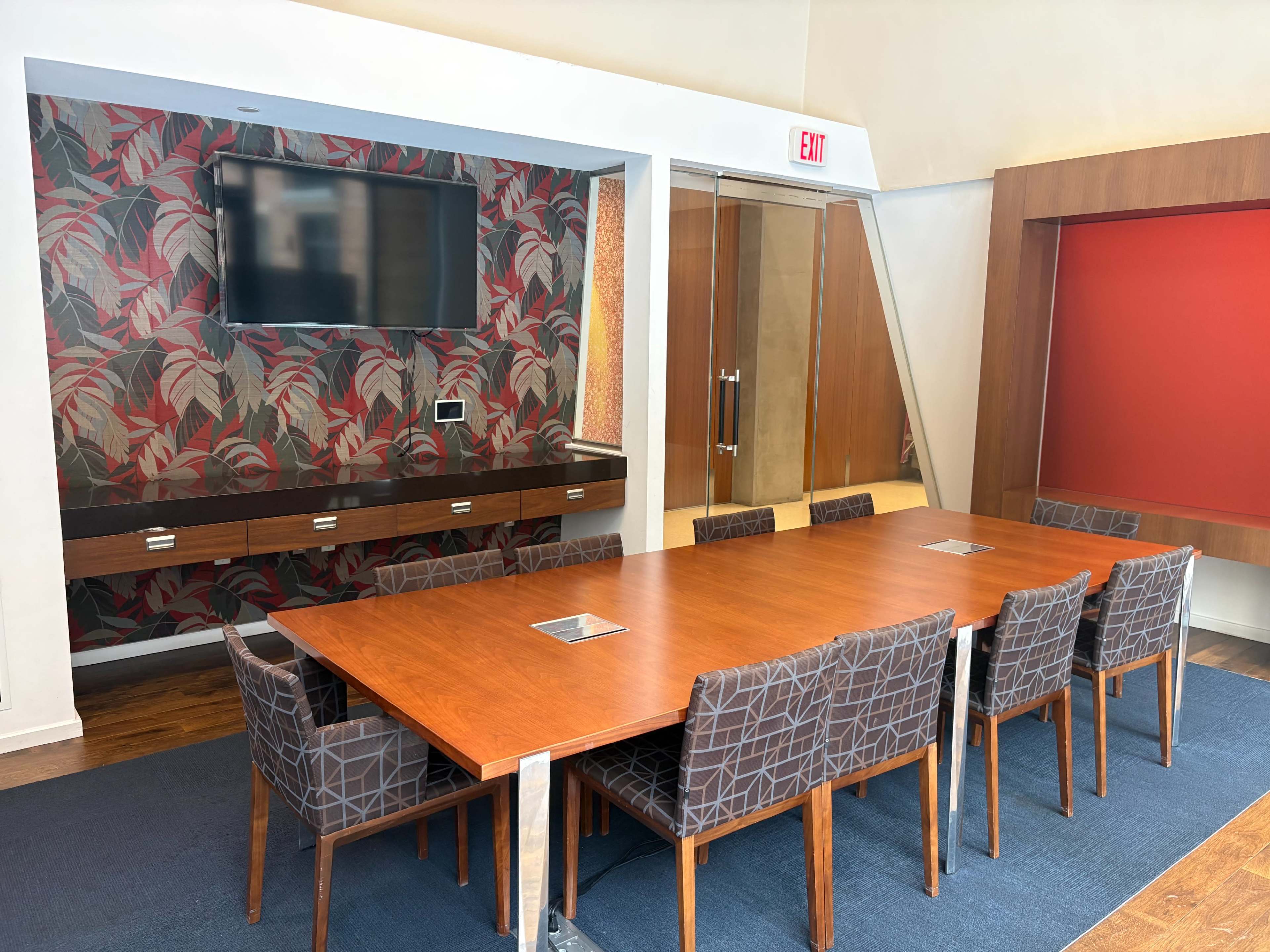 A modern conference room features a large wooden table surrounded by upholstered chairs, with a wall-mounted screen and decorative wallpaper.