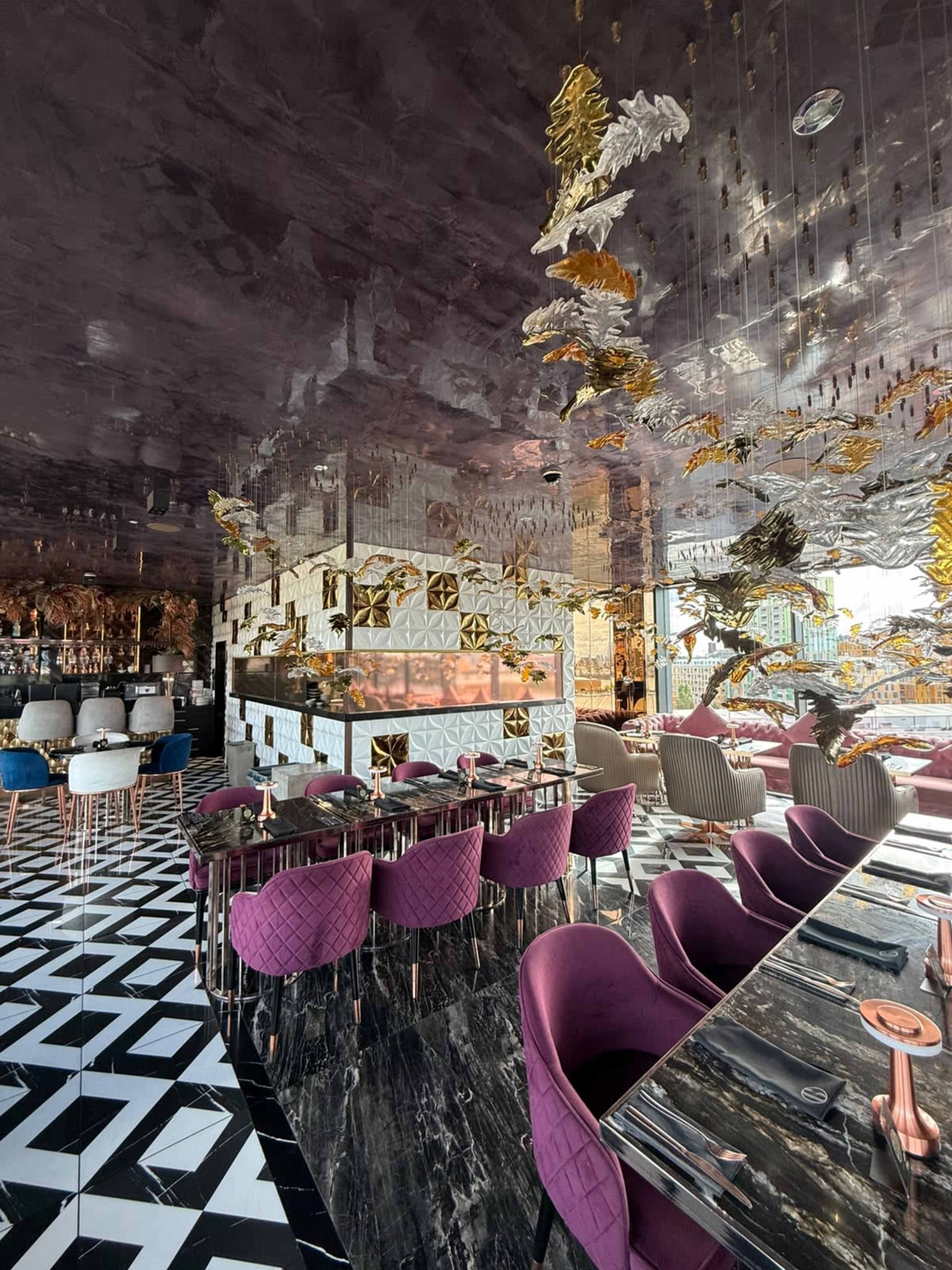 The image shows a stylish restaurant interior featuring a patterned black and white floor, plush purple seating, and a reflective ceiling adorned with decorative elements.