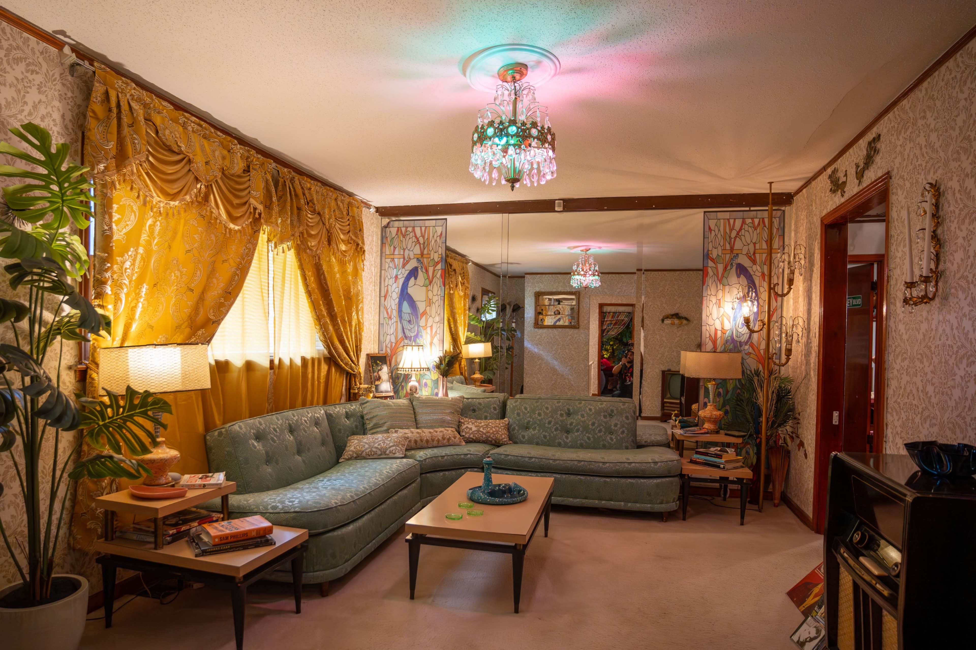 A retro living room features a turquoise sectional couch, patterned walls, and colorful chandeliers providing ambient light.