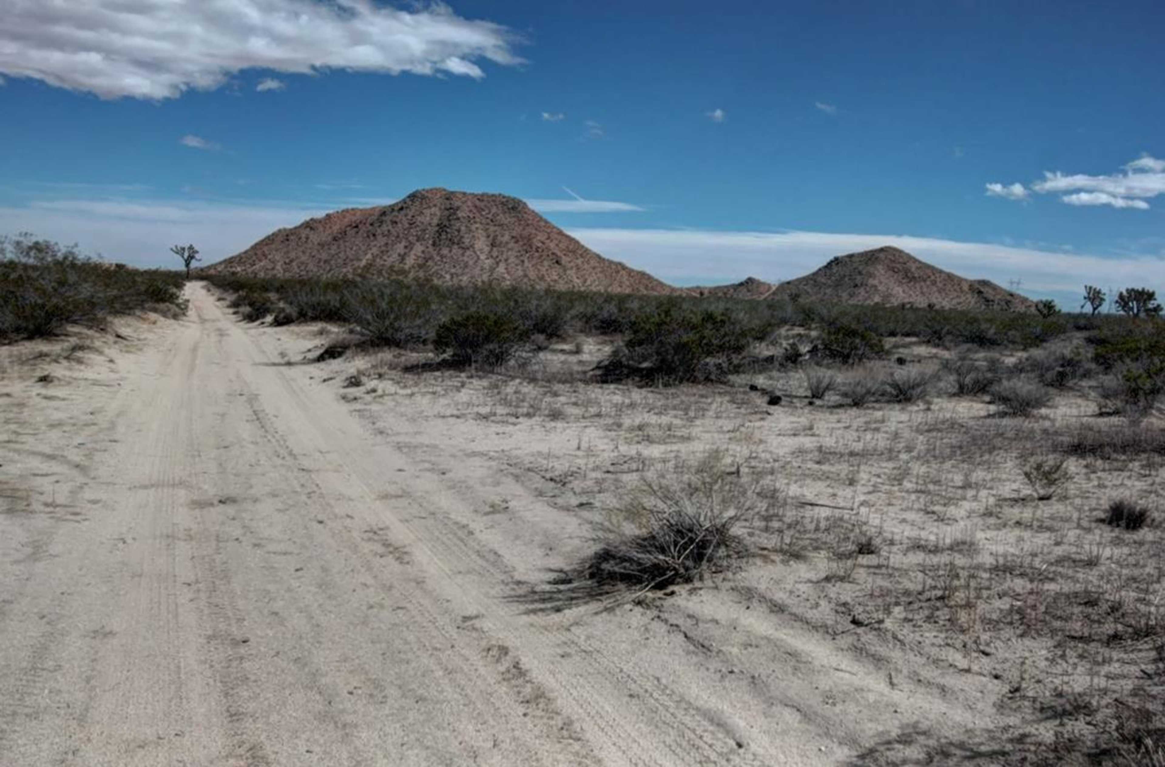 A dirt road leads through a sparse, desert landscape with rocky hills in the distance under a blue sky.