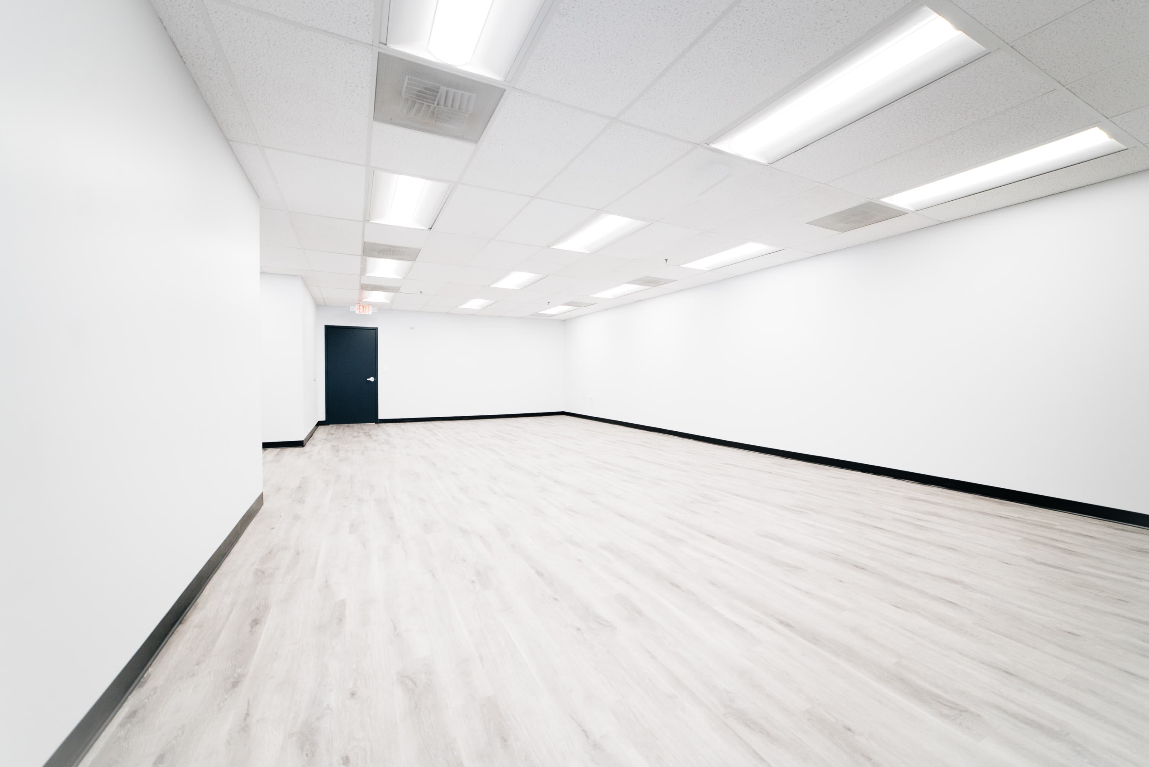 The image shows a spacious, empty room with white walls, a light-colored flooring, and a dark door.