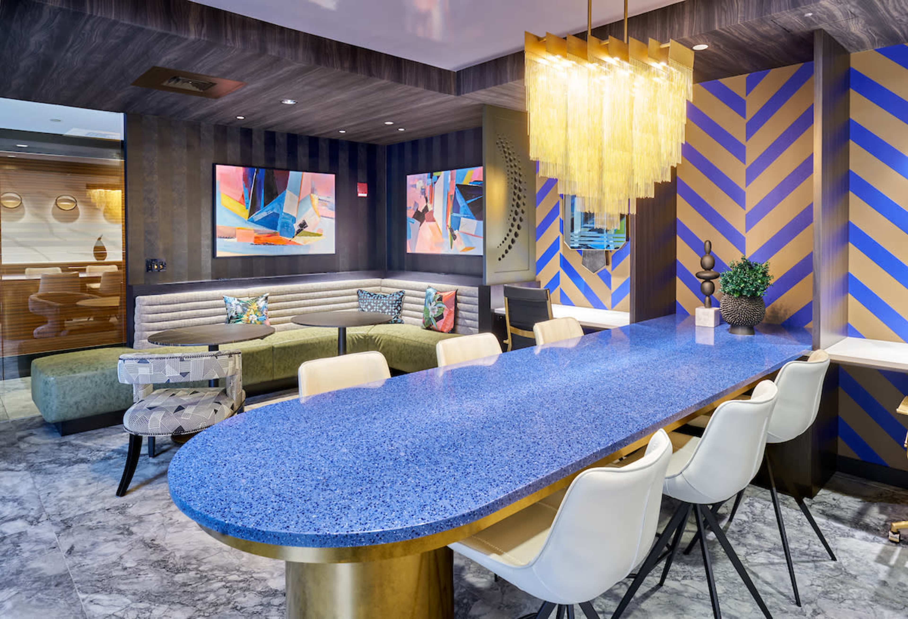 The image shows a modern lounge area with a long blue countertop, white chairs, a green sofa, and colorful artwork on the walls.