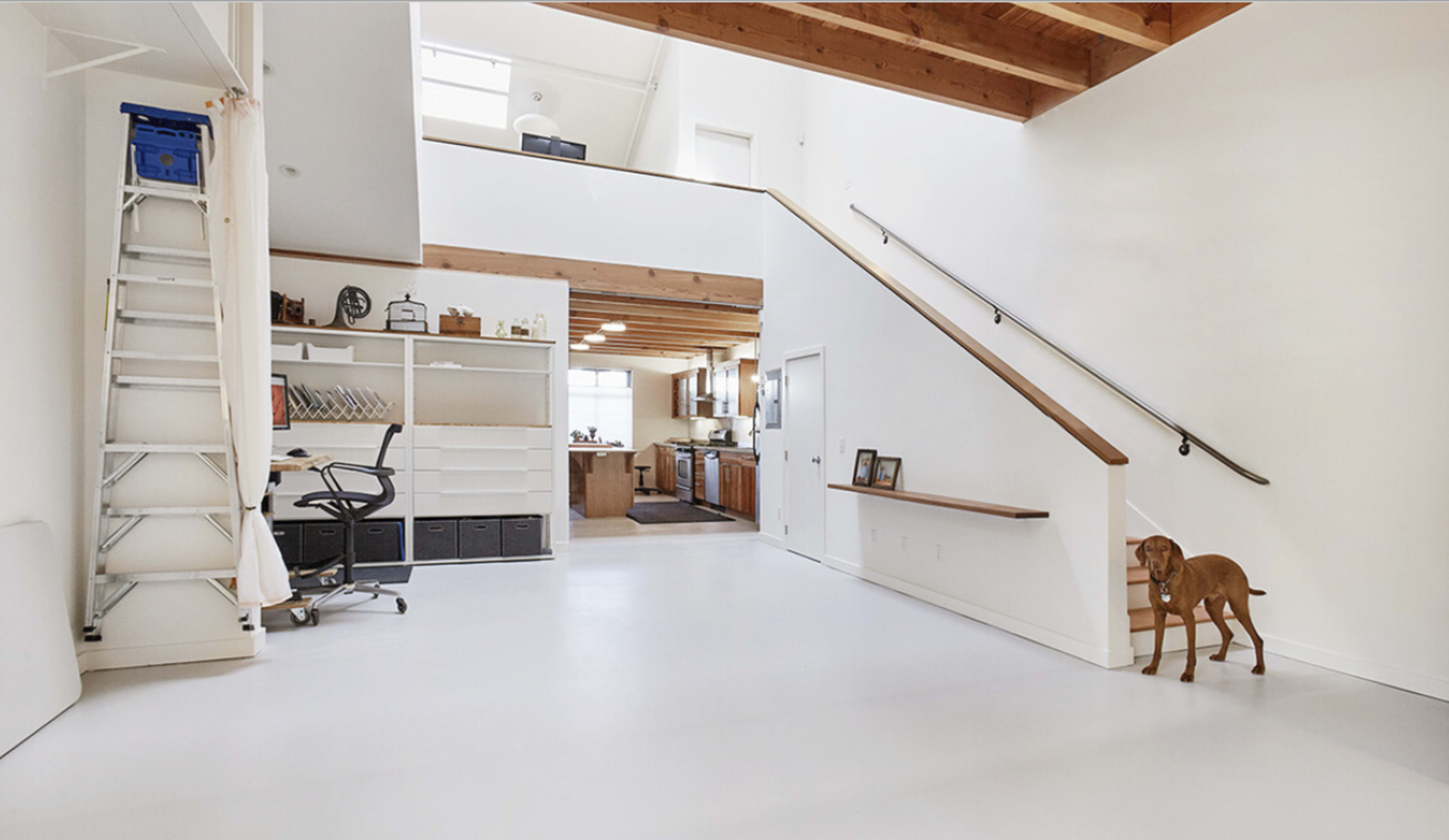 An open-concept interior with a staircase, a desk and chair, a shelving unit, and a dog standing near the stairs.