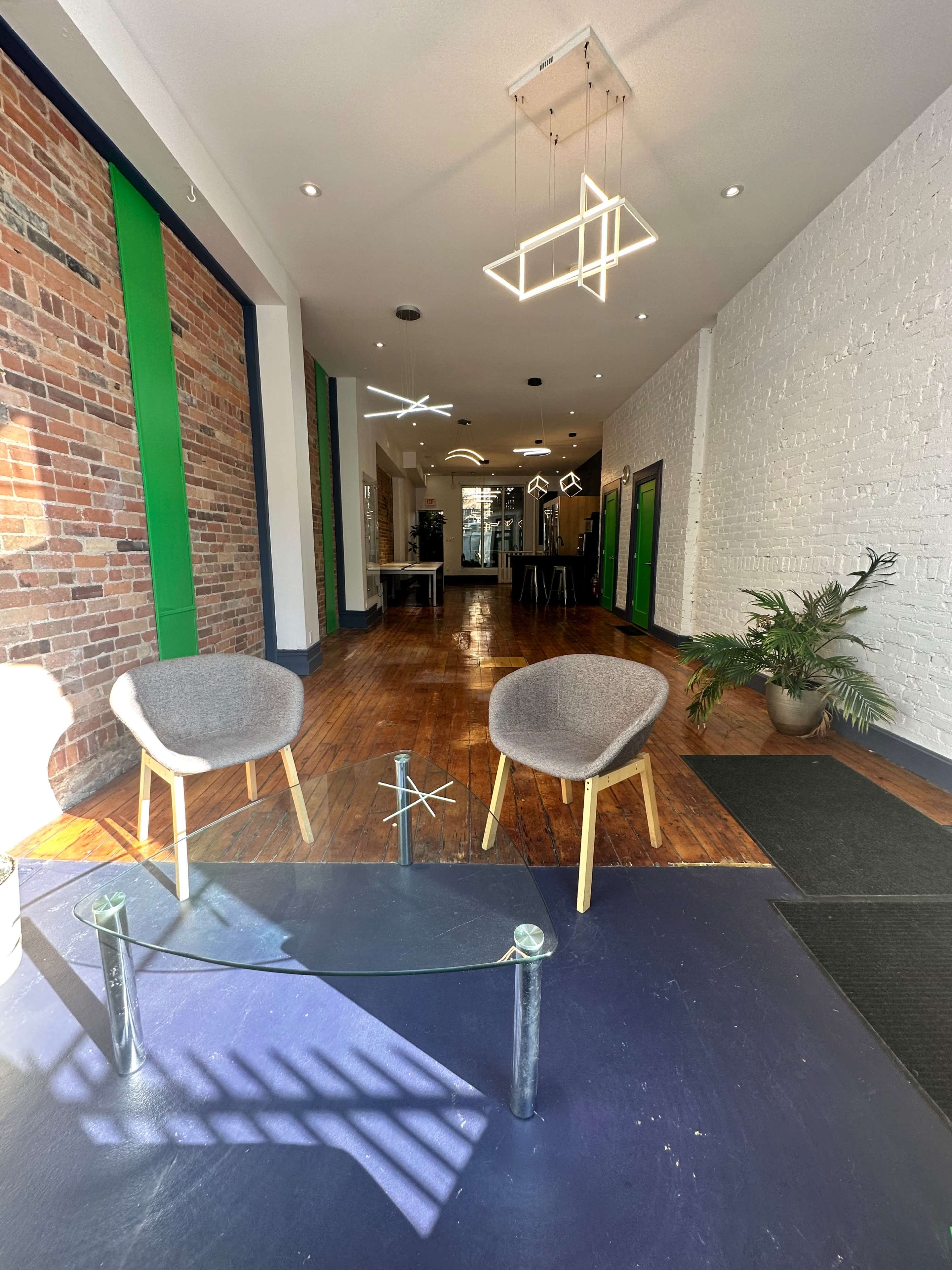 A spacious interior with exposed brick walls, two gray chairs, a glass coffee table, and green accent walls, leading to a well-lit area in the back.