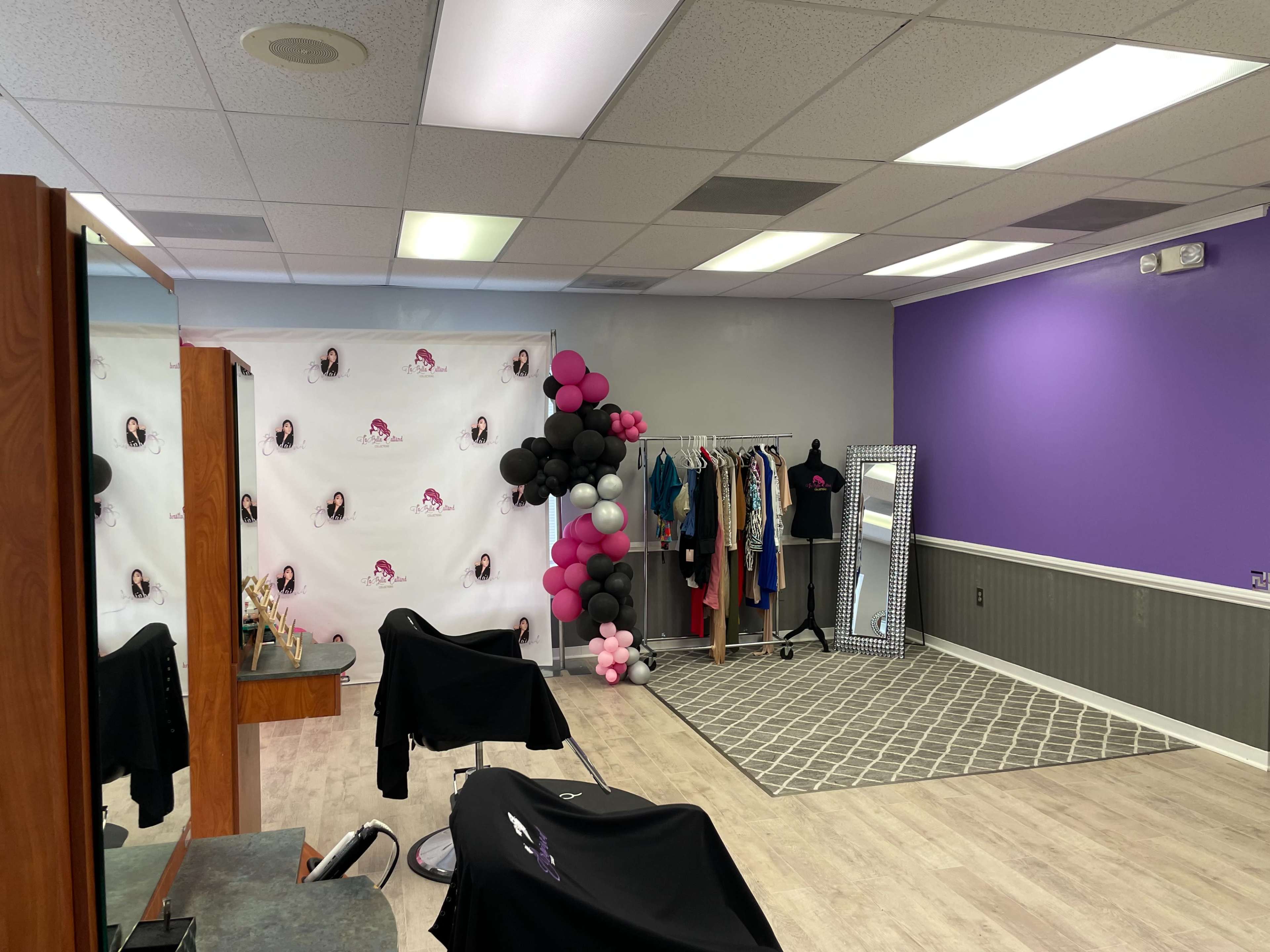 A salon interior featuring styling chairs, a clothing rack with various garments, a large mirror, and a wall decorated with purple paint and balloon arrangements.