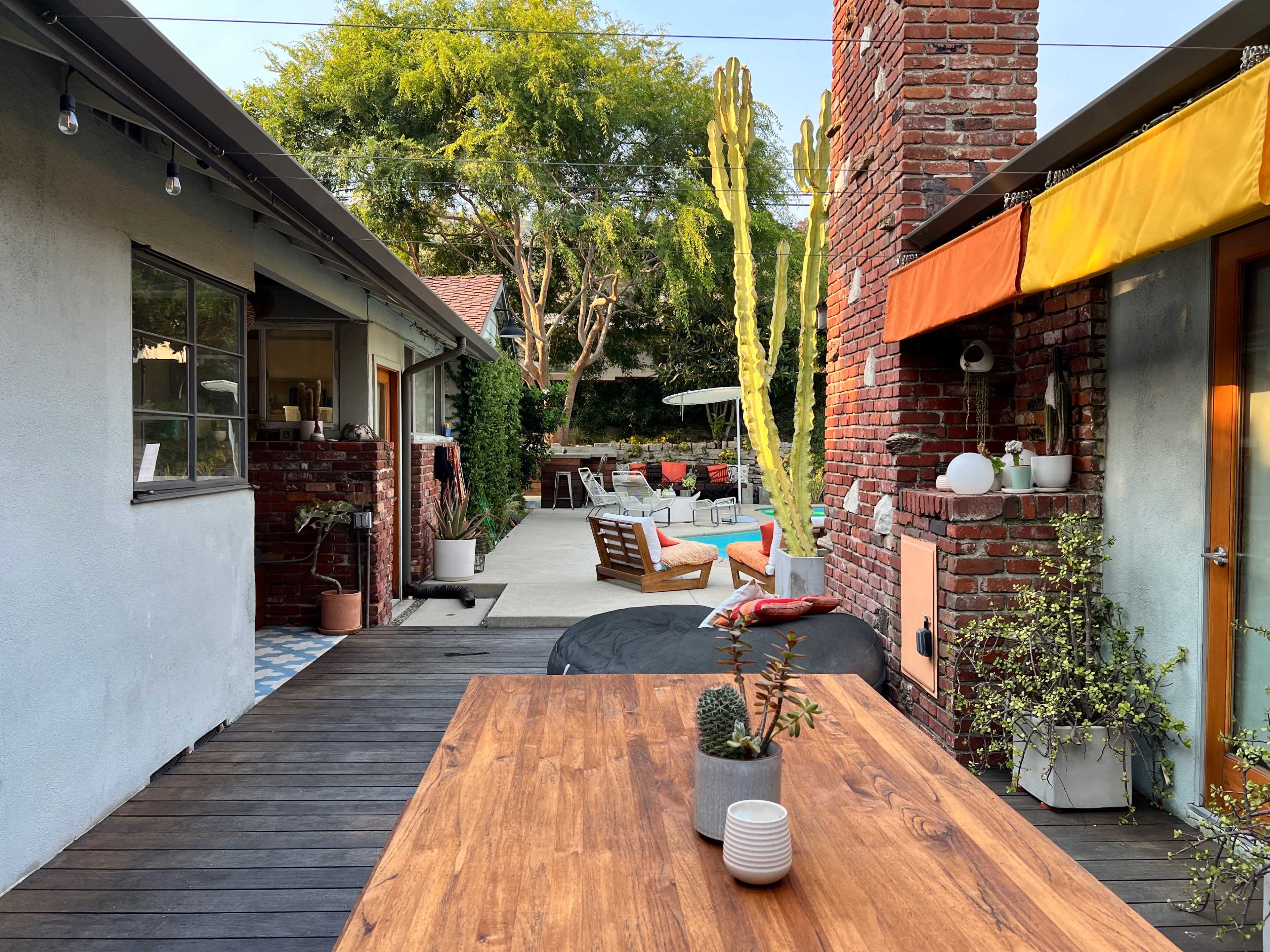 A wooden table with potted plants stands in the foreground, while a backyard with a pool and lounge chairs is visible in the background, surrounded by brick walls and greenery.