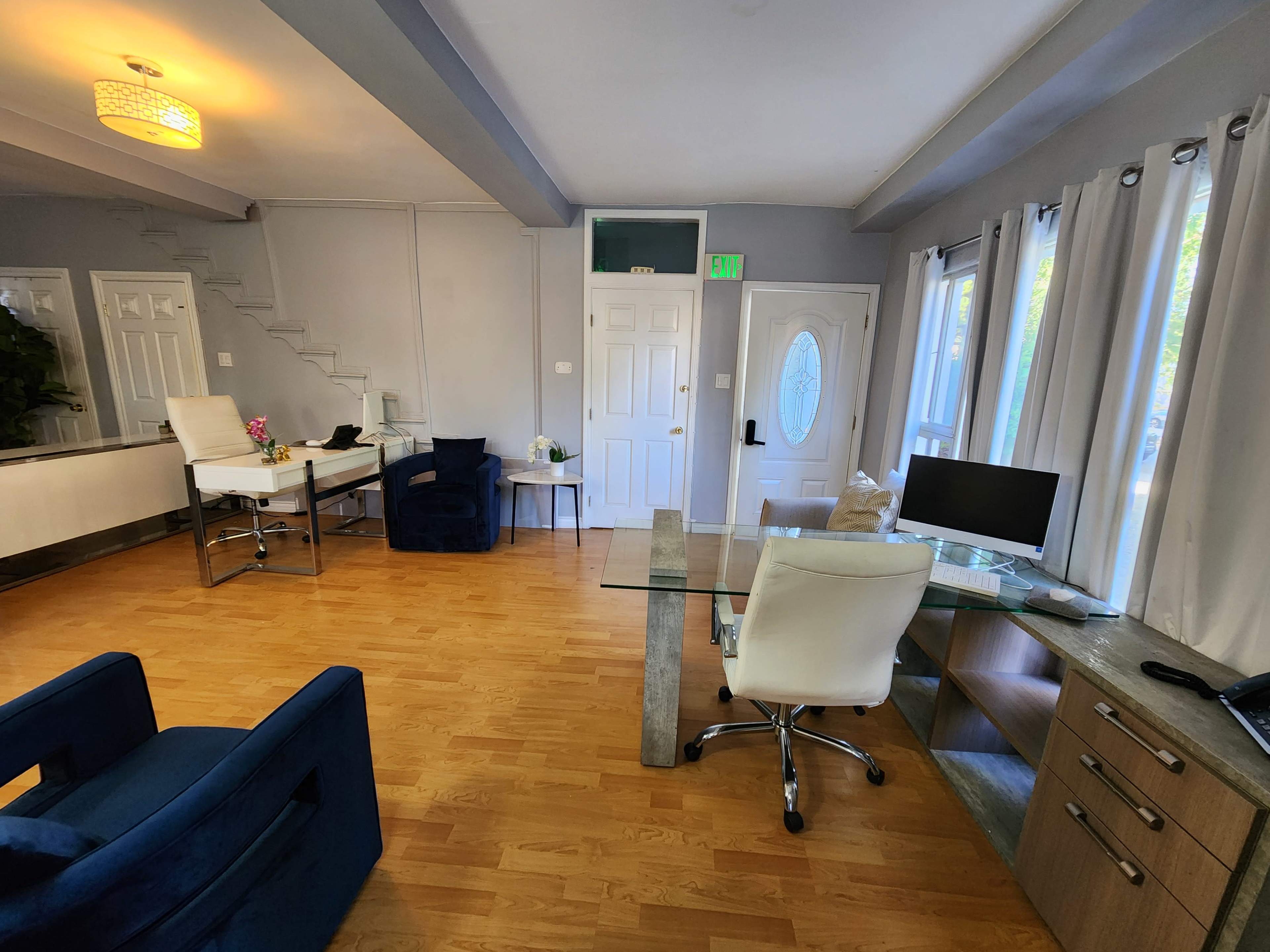 The image shows a modern, well-lit living space featuring a combination of office furniture and a seating area with a staircase in the background.