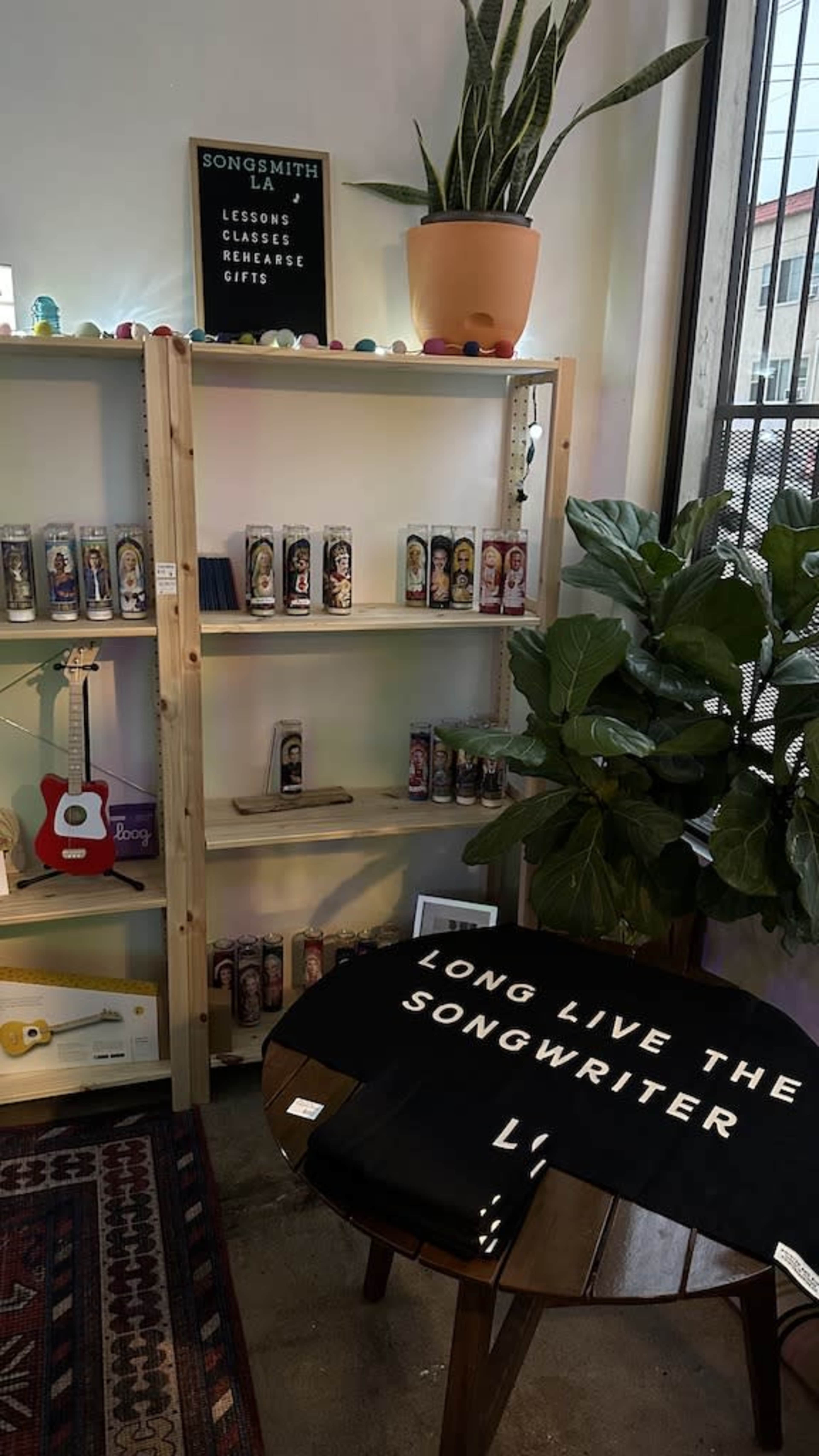 A wooden shelf displays colorful candles and a small guitar next to a potted plant, with a black shirt featuring the text "LONG LIVE THE SONGWRITER" draped across a table.