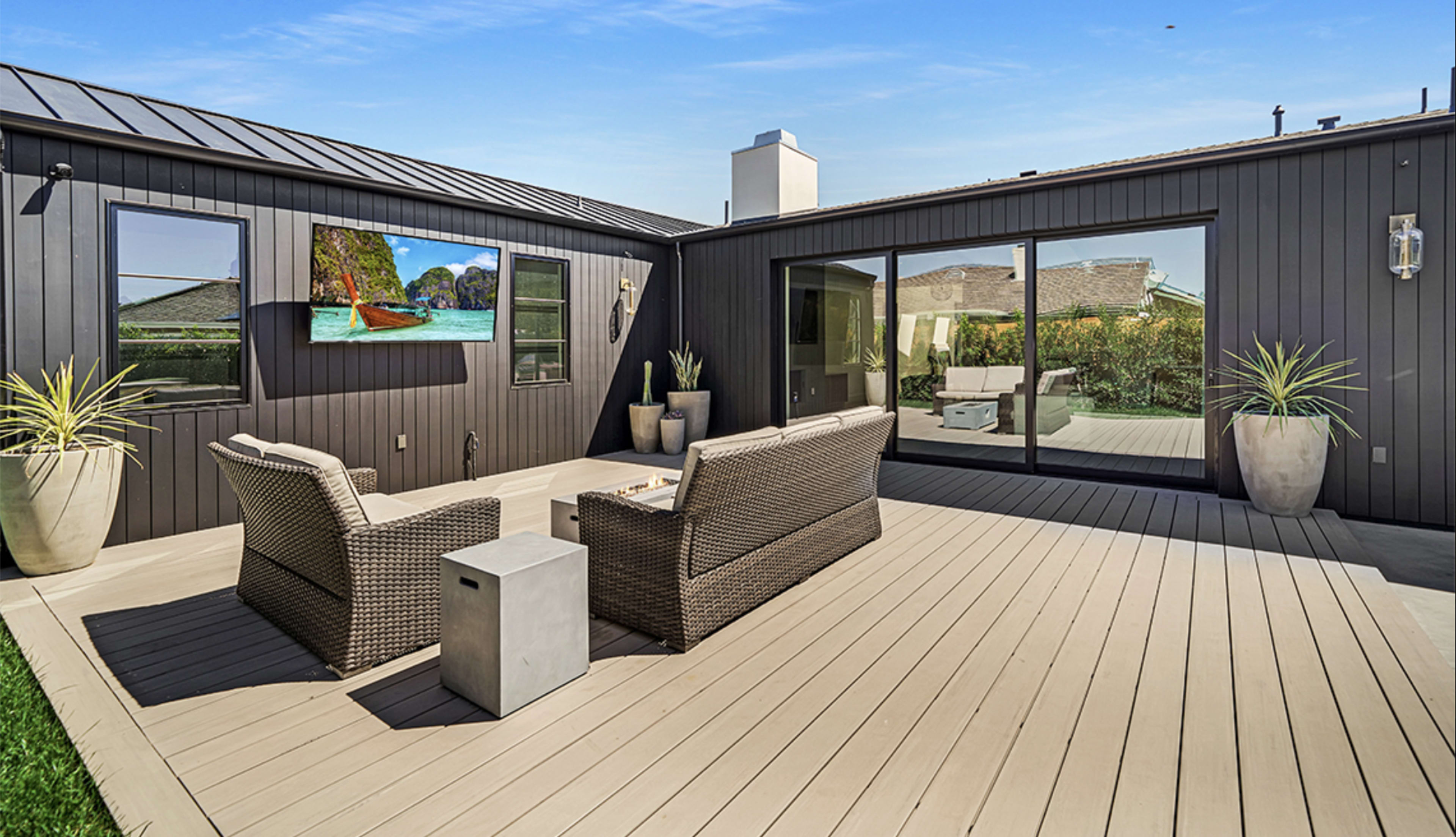 The image shows a modern outdoor patio with wicker furniture, a television mounted on the wall, and sliding glass doors leading into a house.