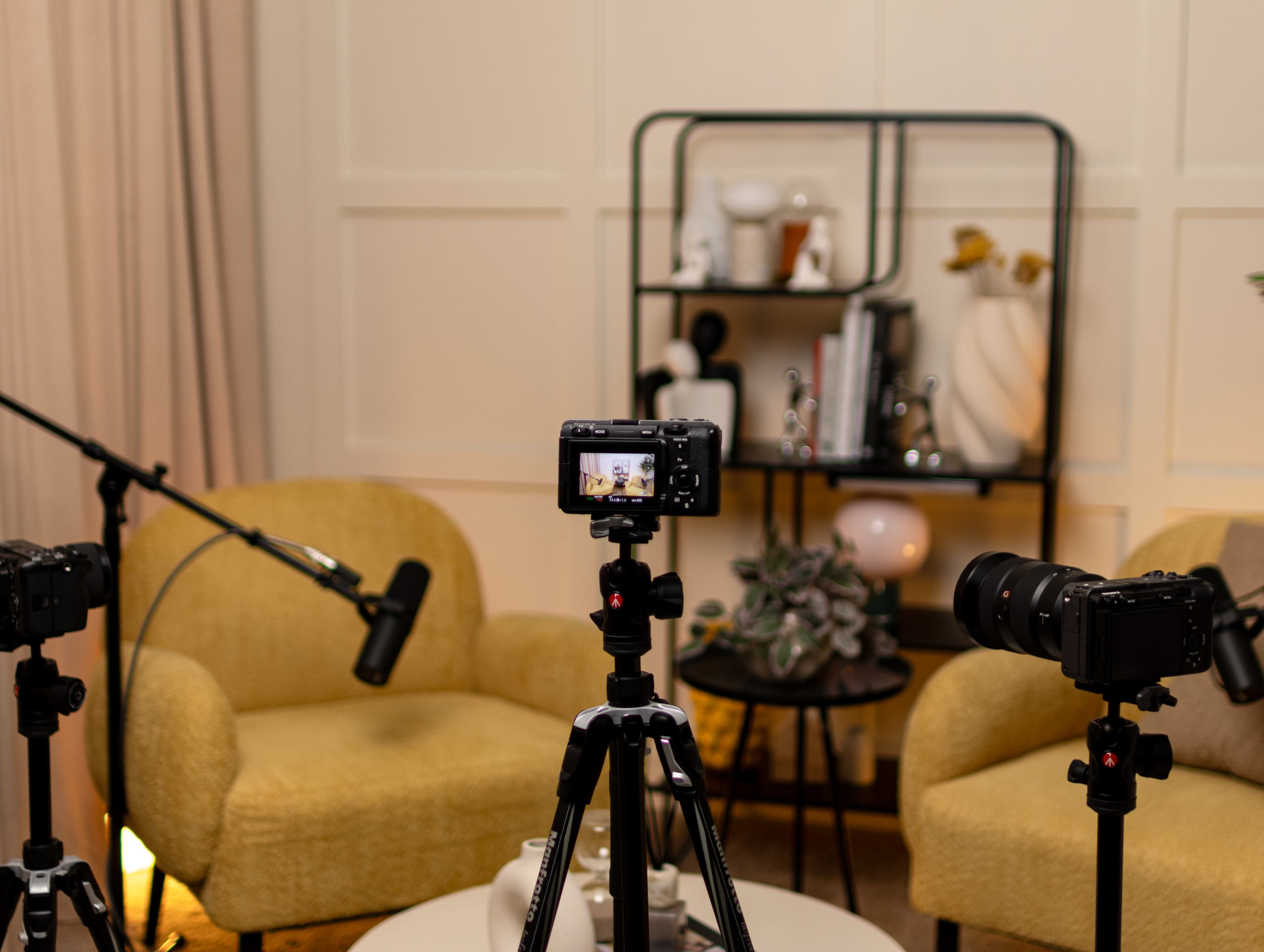 The image shows a setup for a video recording session featuring two yellow armchairs, tripods with cameras, and a microphone.