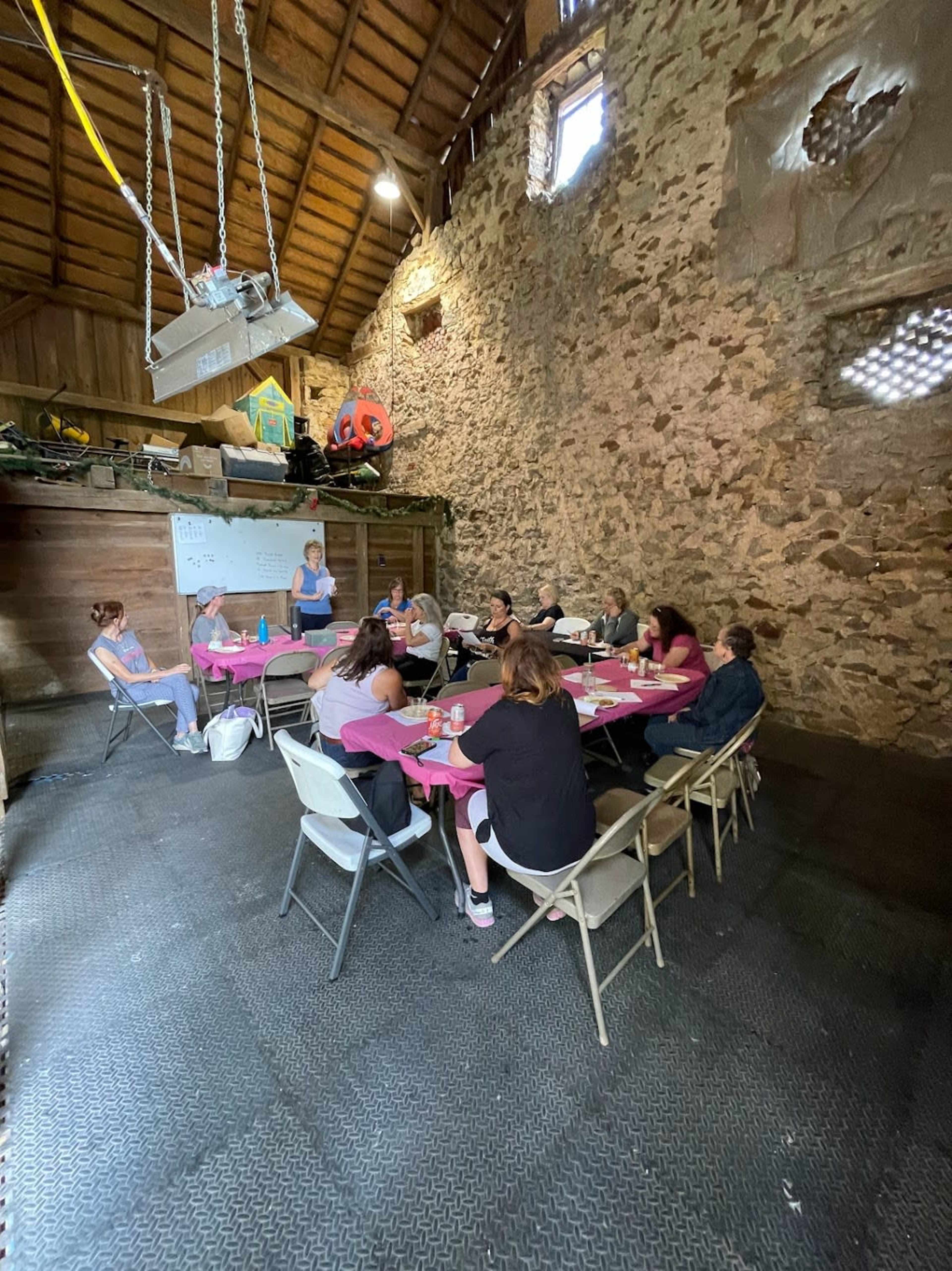 A group of people sits around tables covered with pink tablecloths in a rustic stone building while a woman stands and presents to them.
