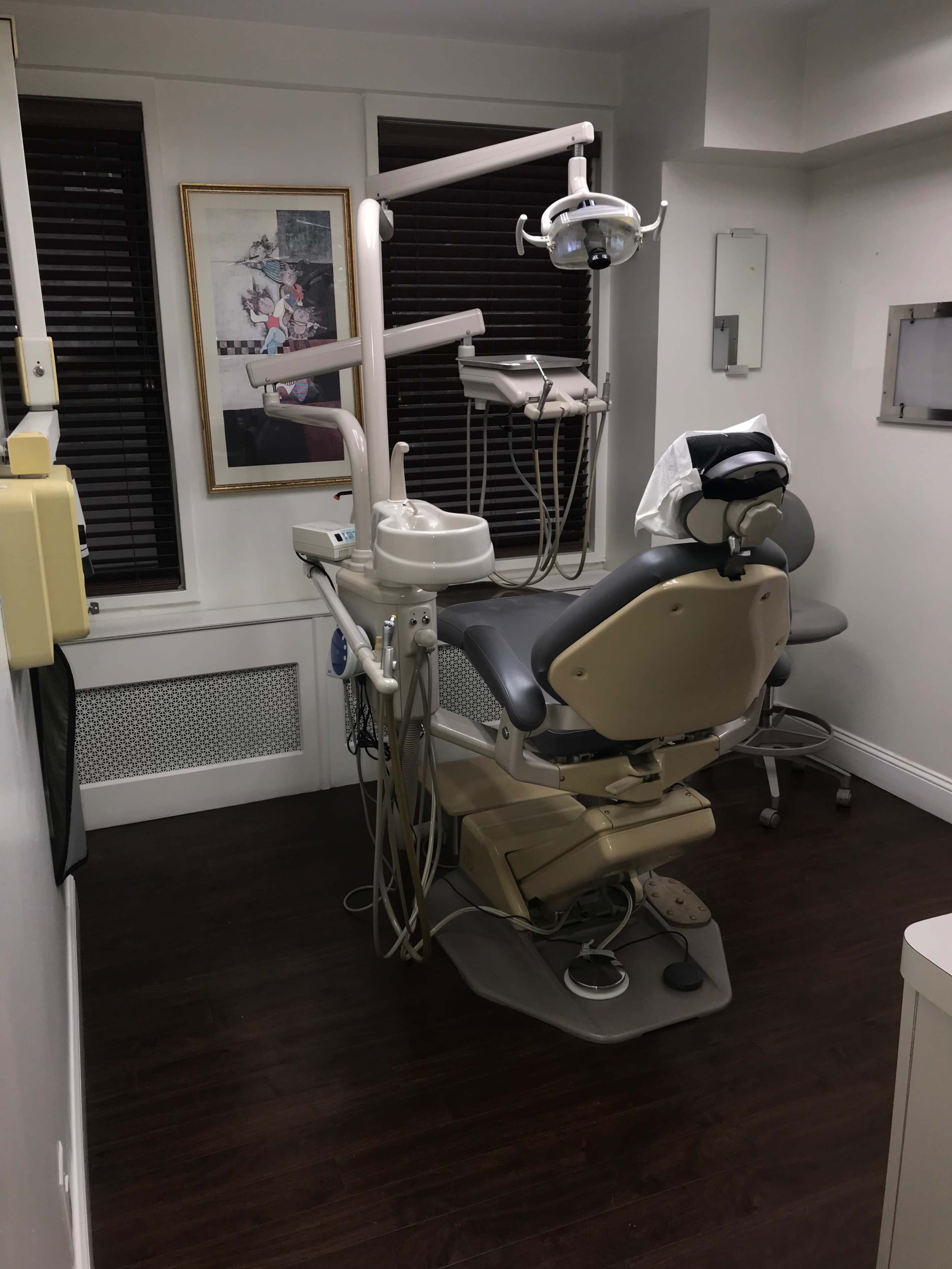 The image shows a dental office with a treatment chair, dental equipment, and a framed picture on the wall.