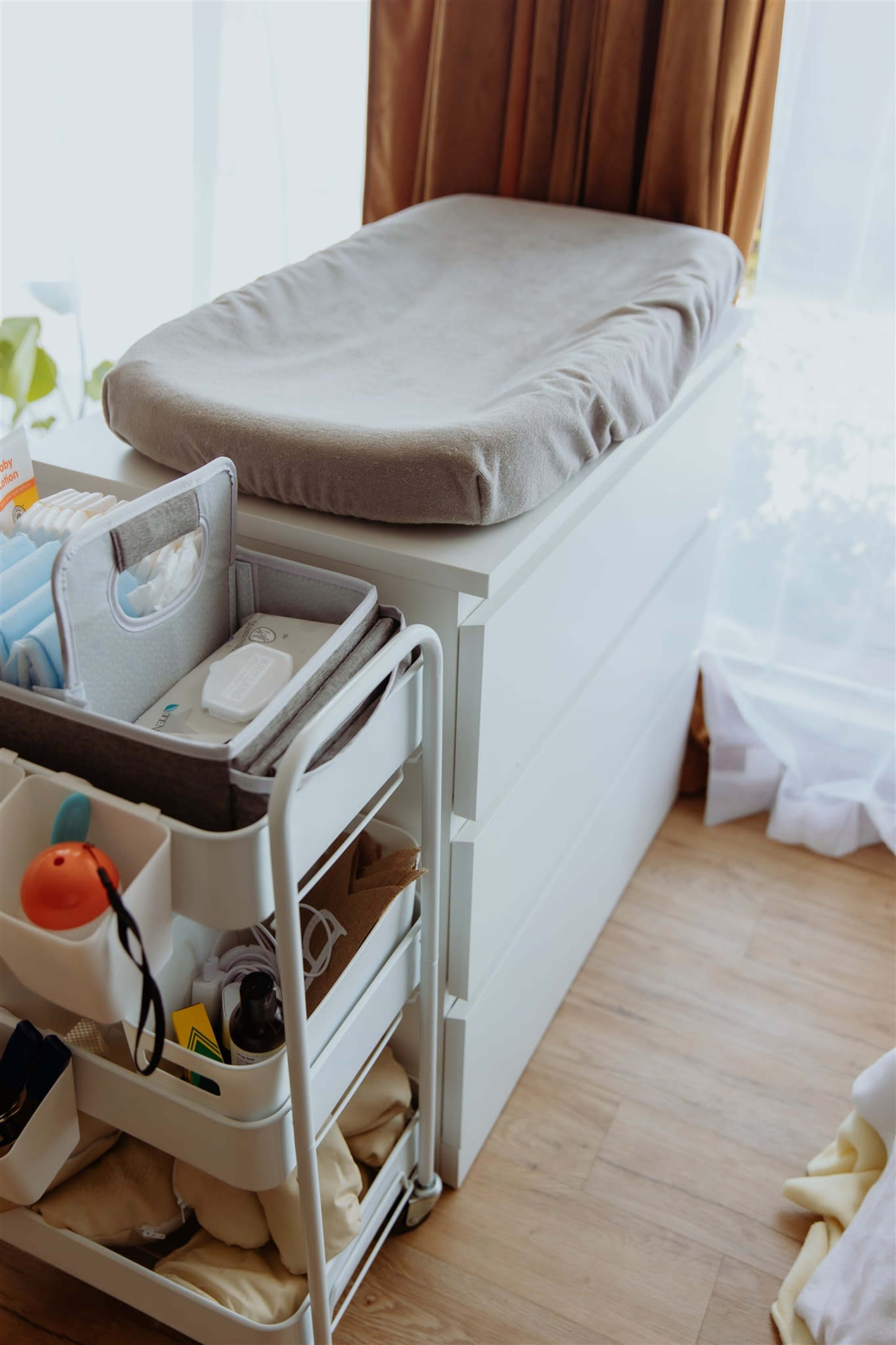 A white dresser with a changing pad on top is adjacent to a cart filled with baby supplies and storage containers.