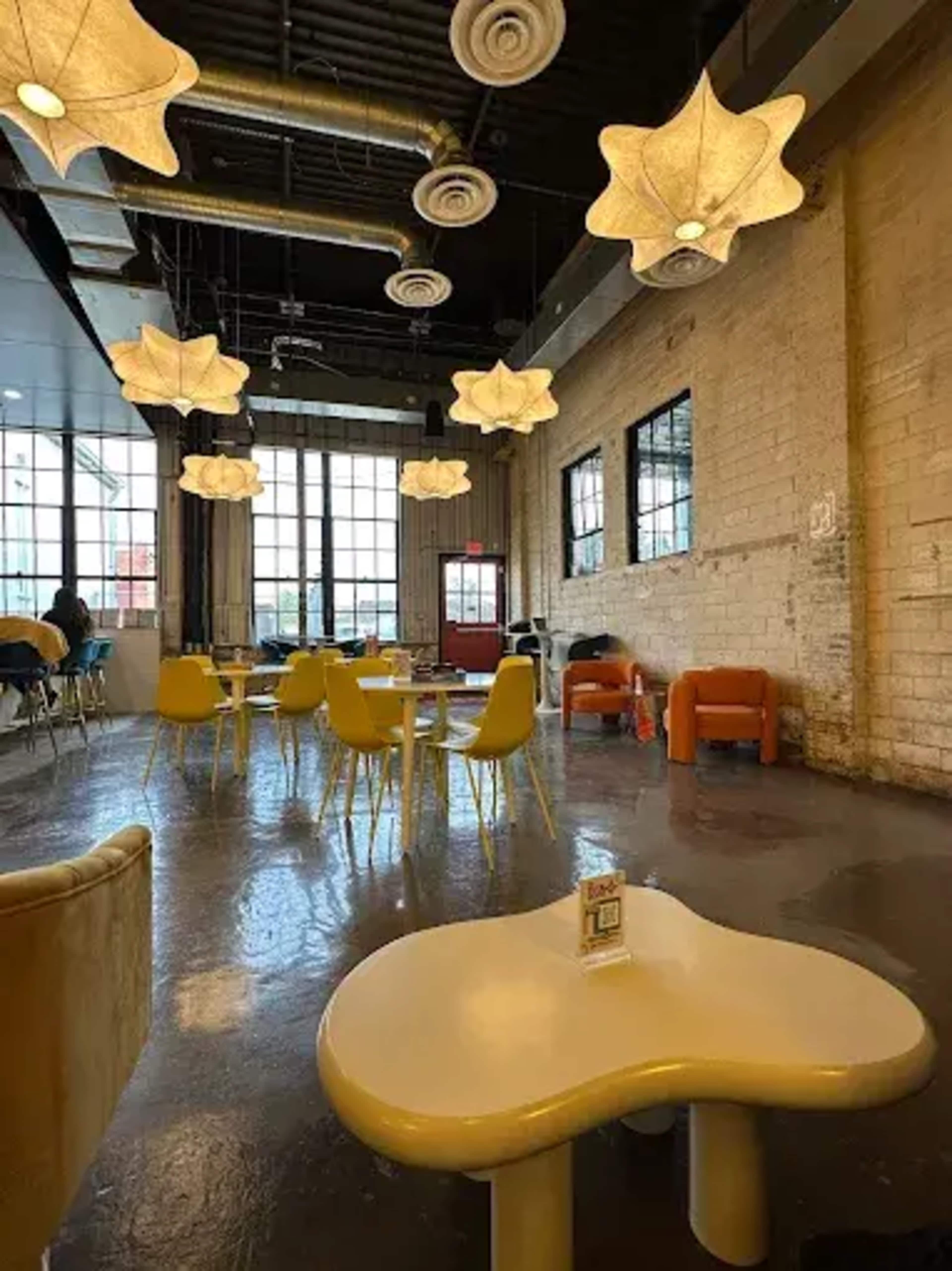 The image shows a spacious café with unique star-shaped light fixtures, colorful chairs, and a large window allowing natural light to fill the room.