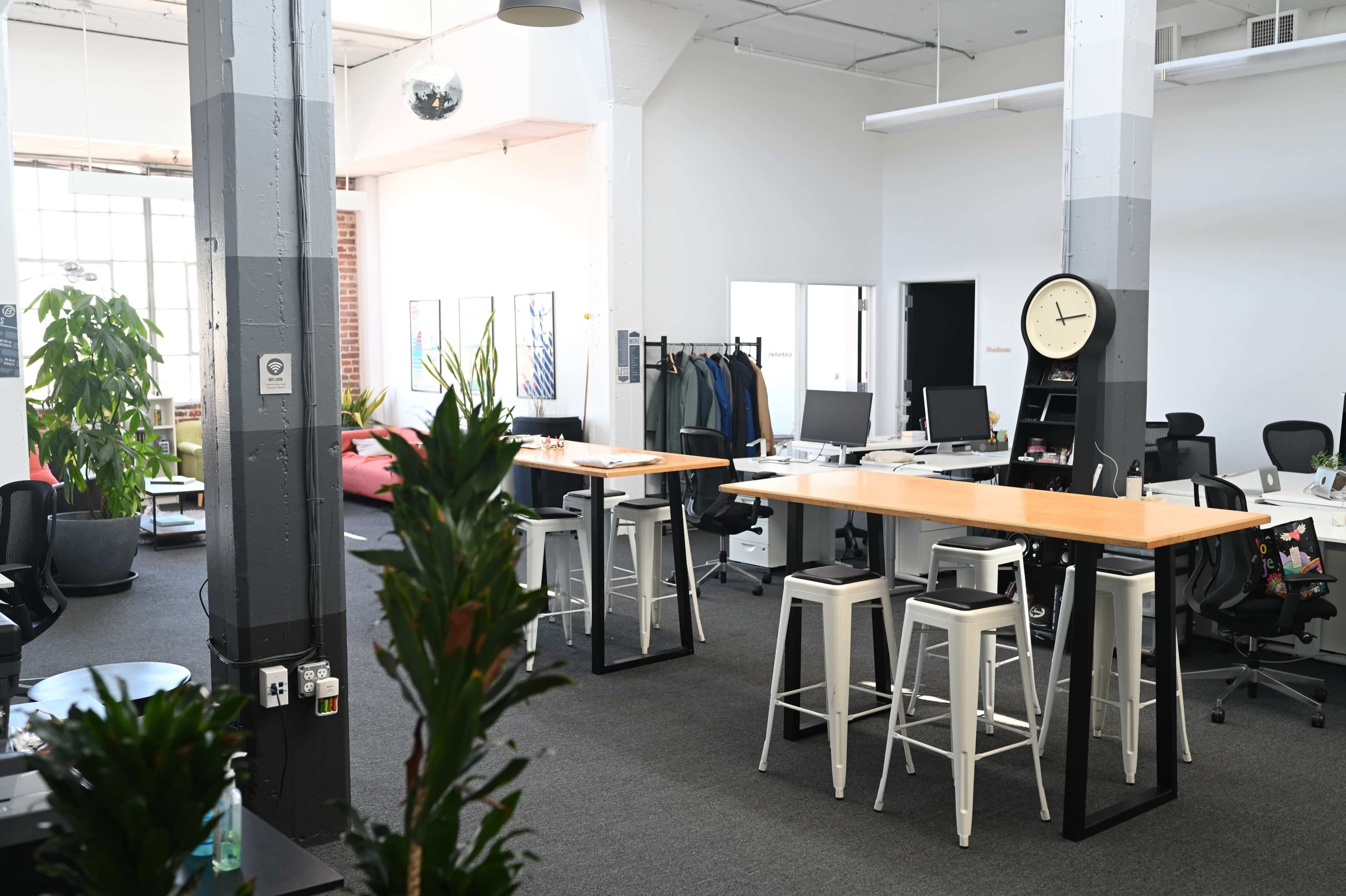 The image shows a modern office space with tall tables, bar stools, and various workstations, surrounded by plants and organized furniture.