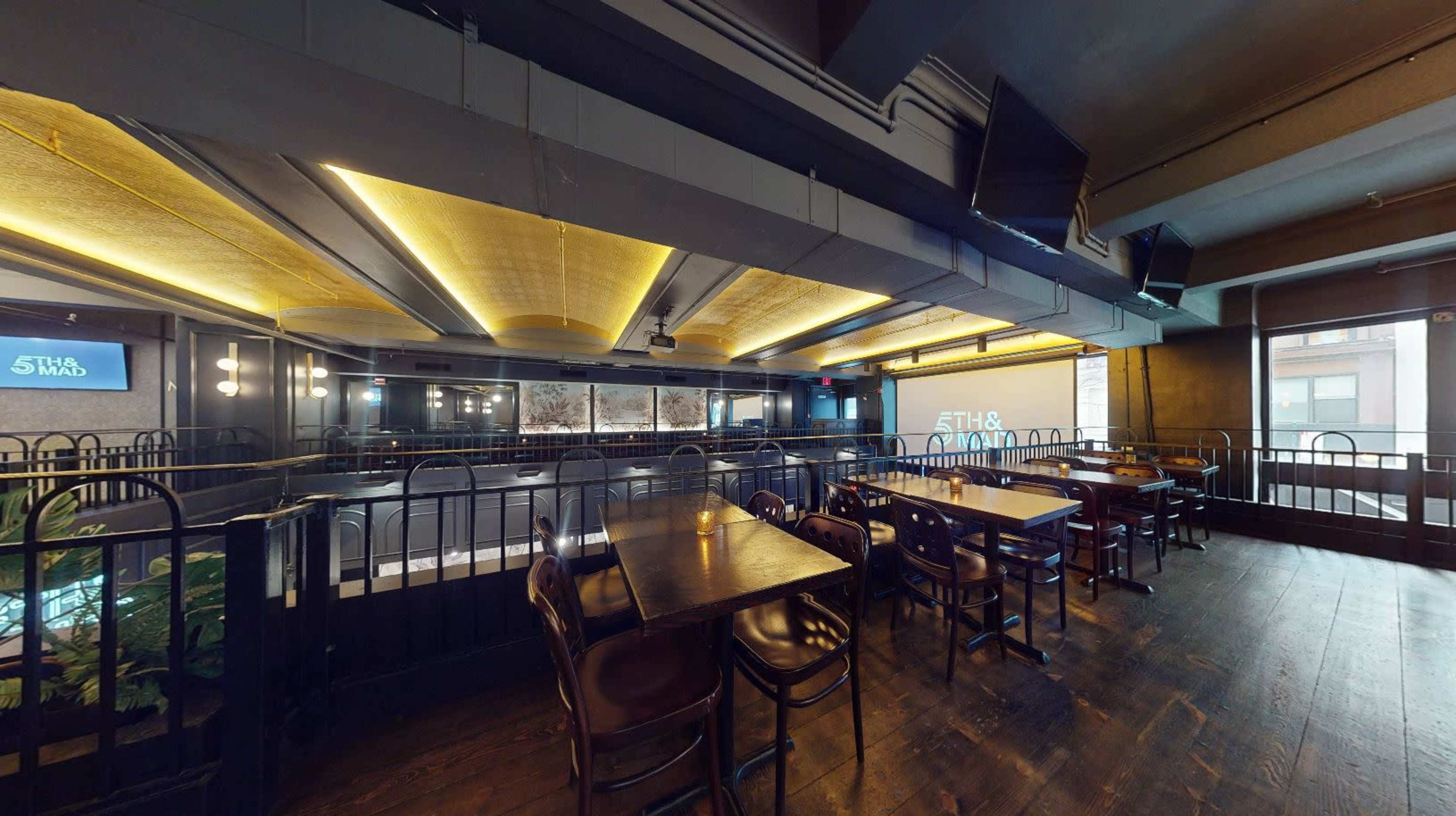 The image shows an interior view of a restaurant with wooden tables and chairs, dim lighting, and a large screen on one wall.