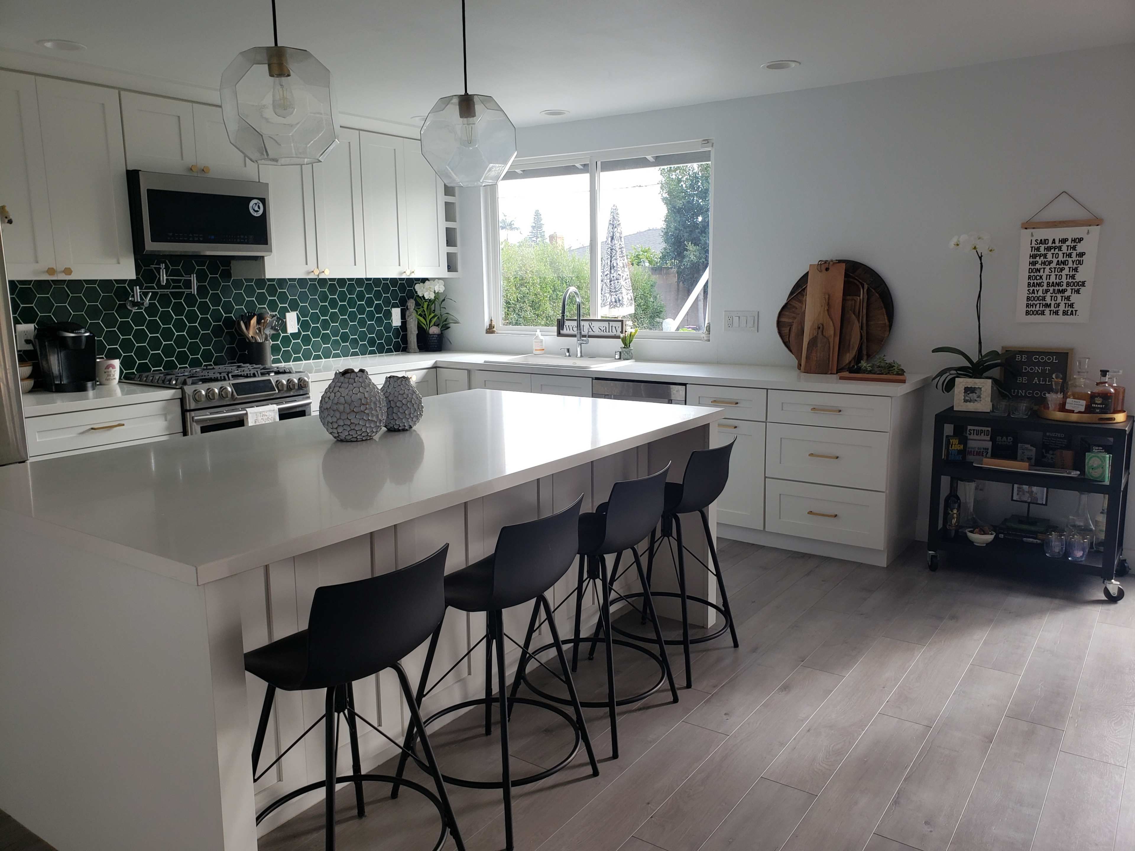 The image shows a modern kitchen featuring white cabinets, a green hexagonal tile backsplash, an island with bar stools, and a large window that allows natural light.