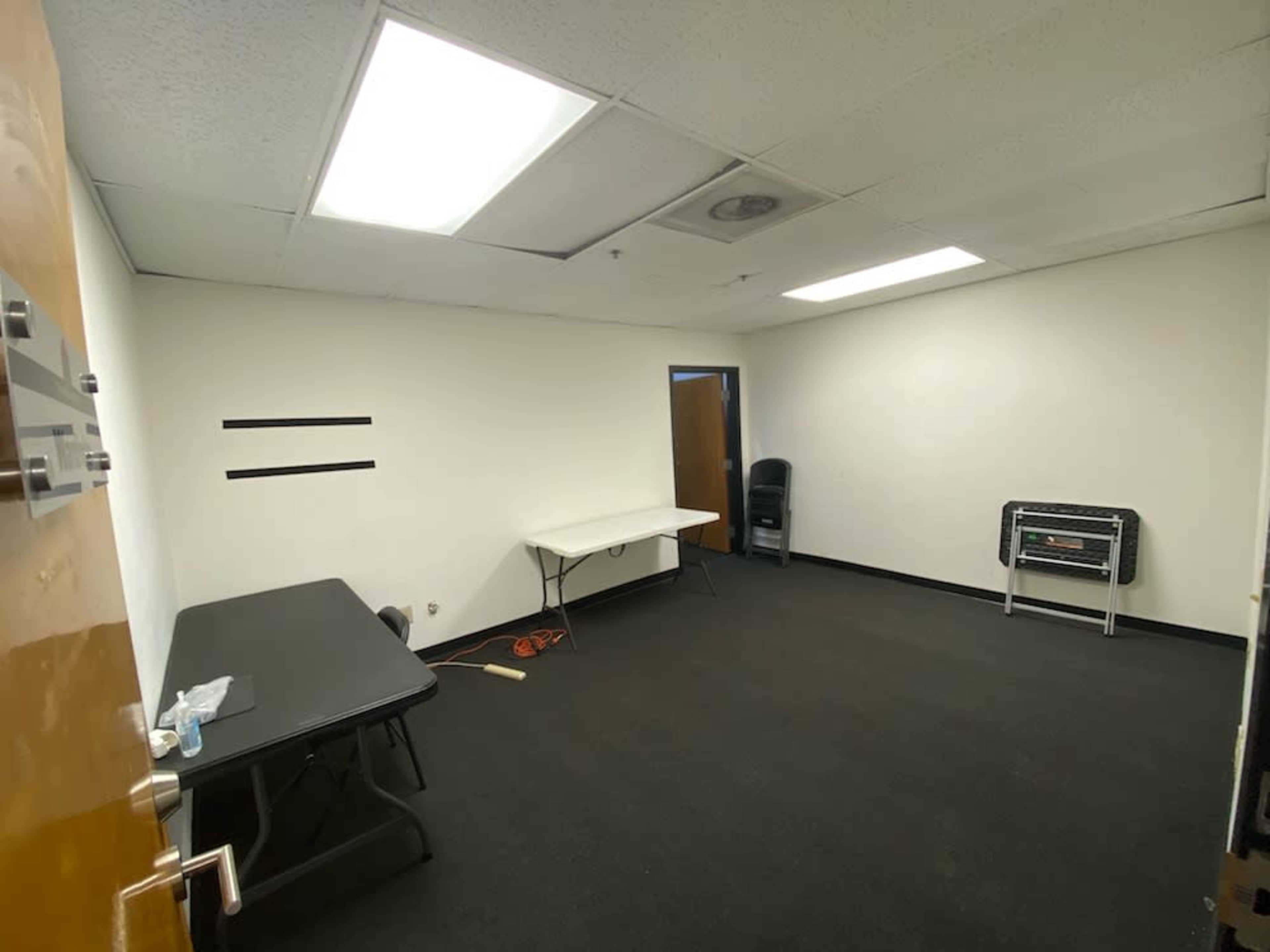 The image shows an empty office room with two folding tables, a chair, and a heater against a plain white wall.