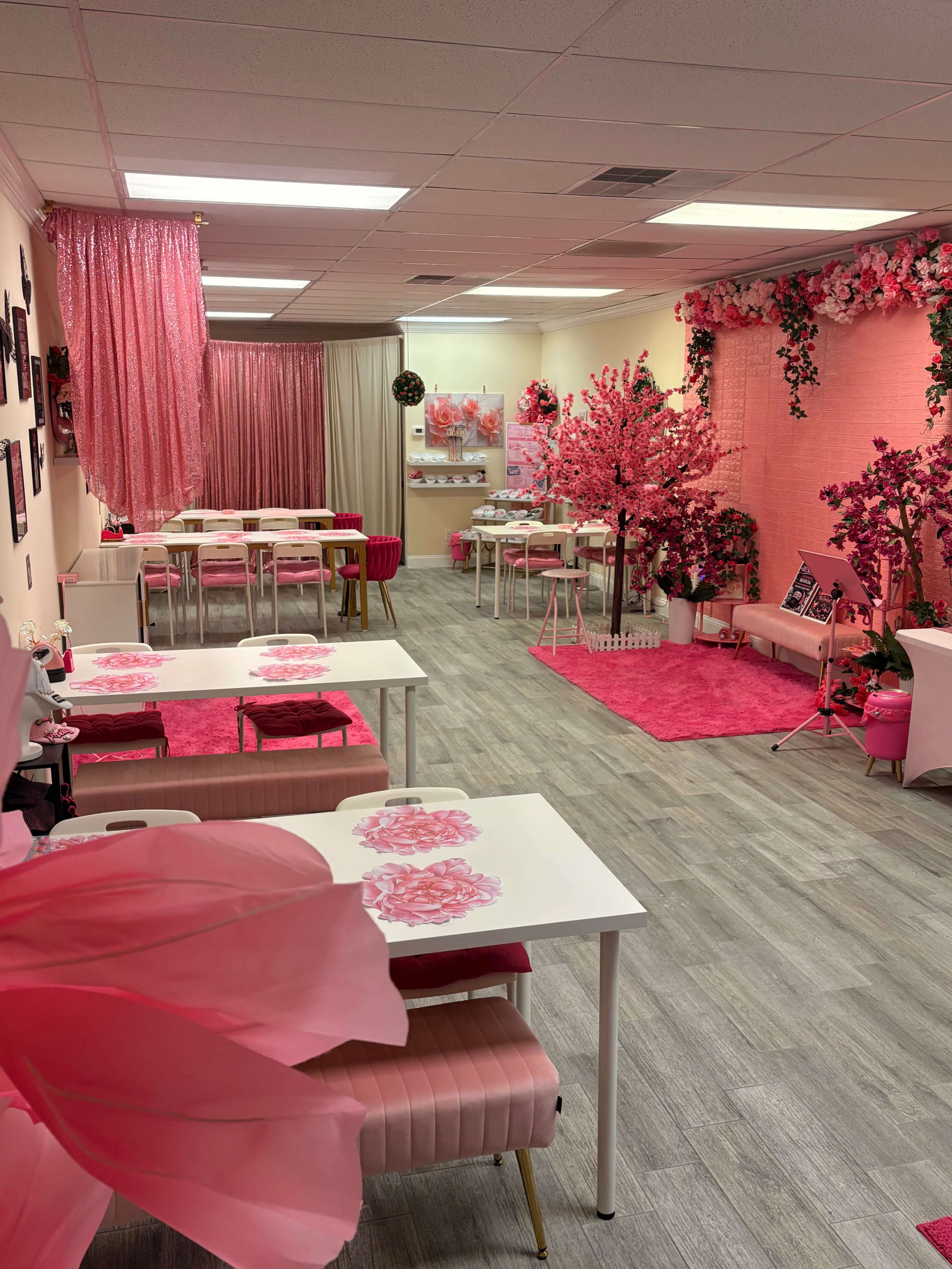 The image shows a brightly decorated room featuring pink-themed furniture, floral arrangements, and multiple tables set up for an event.
