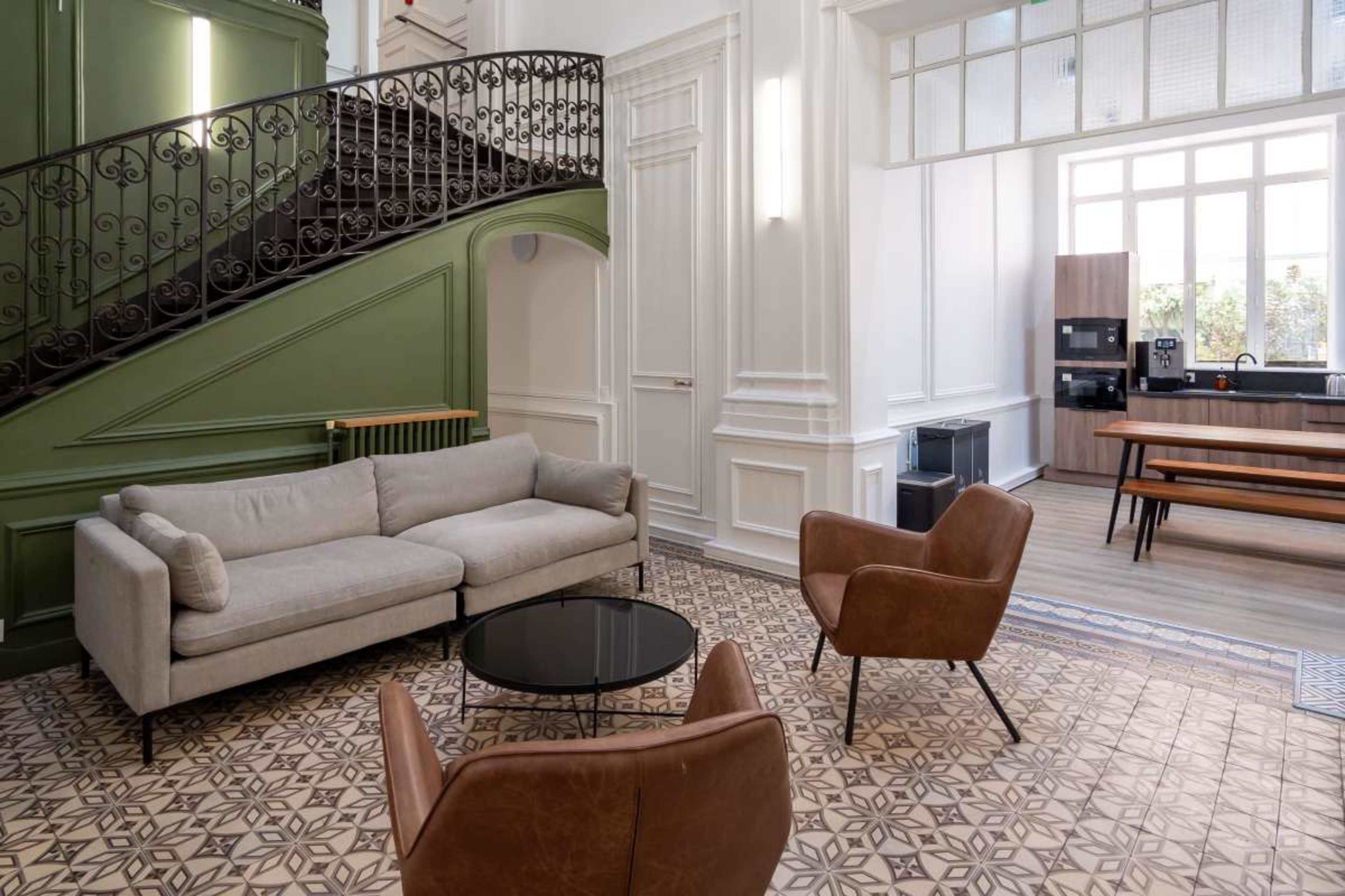 The image shows a stylish living space featuring a gray sofa, two brown chairs, a round black coffee table, and a staircase with intricate wrought iron railings, all set against patterned tiled flooring.