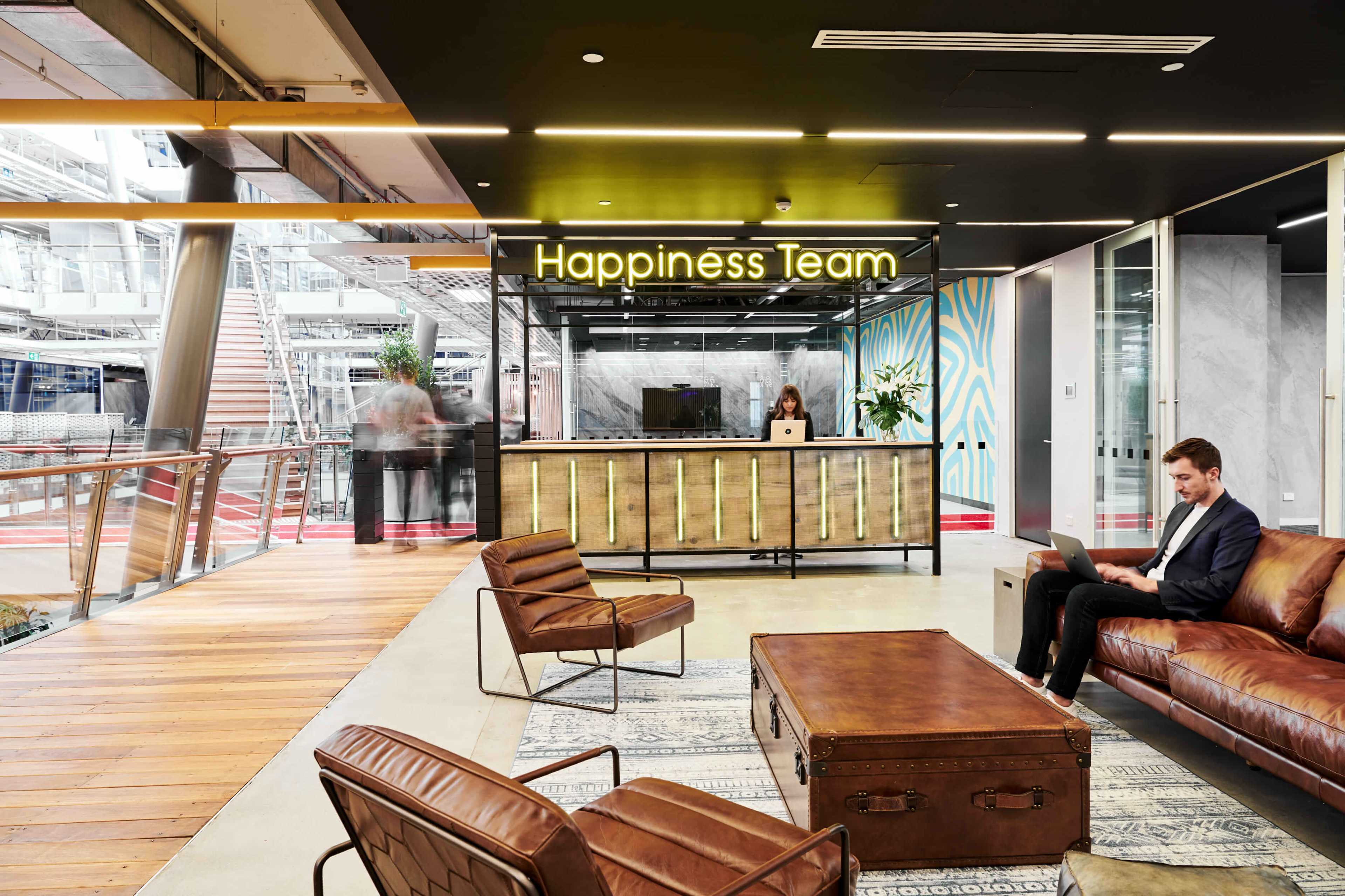 The image shows a modern office reception area with a "Happiness Team" sign, a front desk, a seating area with a leather couch and a coffee table, and a person working on a laptop.