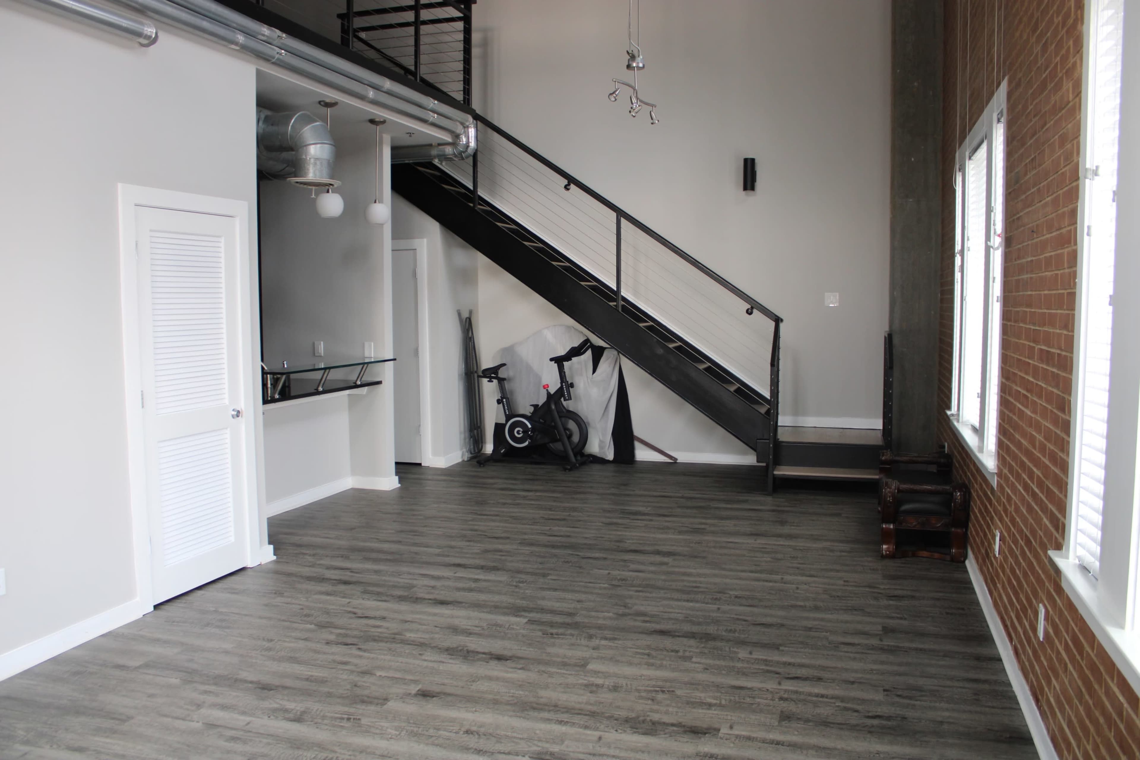 An empty industrial-style living space featuring a staircase, large windows, and a corner with exercise equipment.