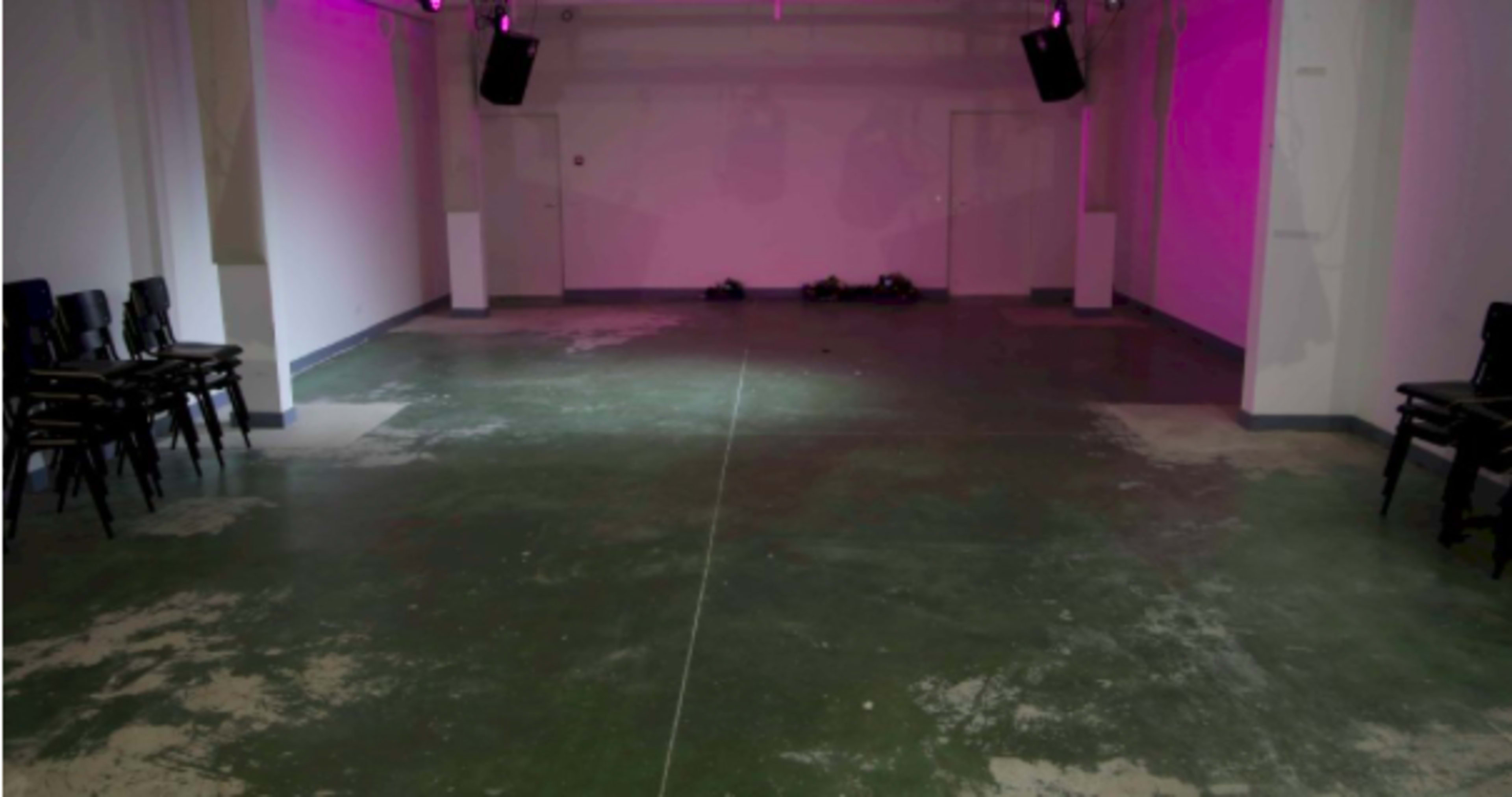 An empty, sparsely furnished room with green flooring, a few stacked chairs against one wall, and ambient lighting.