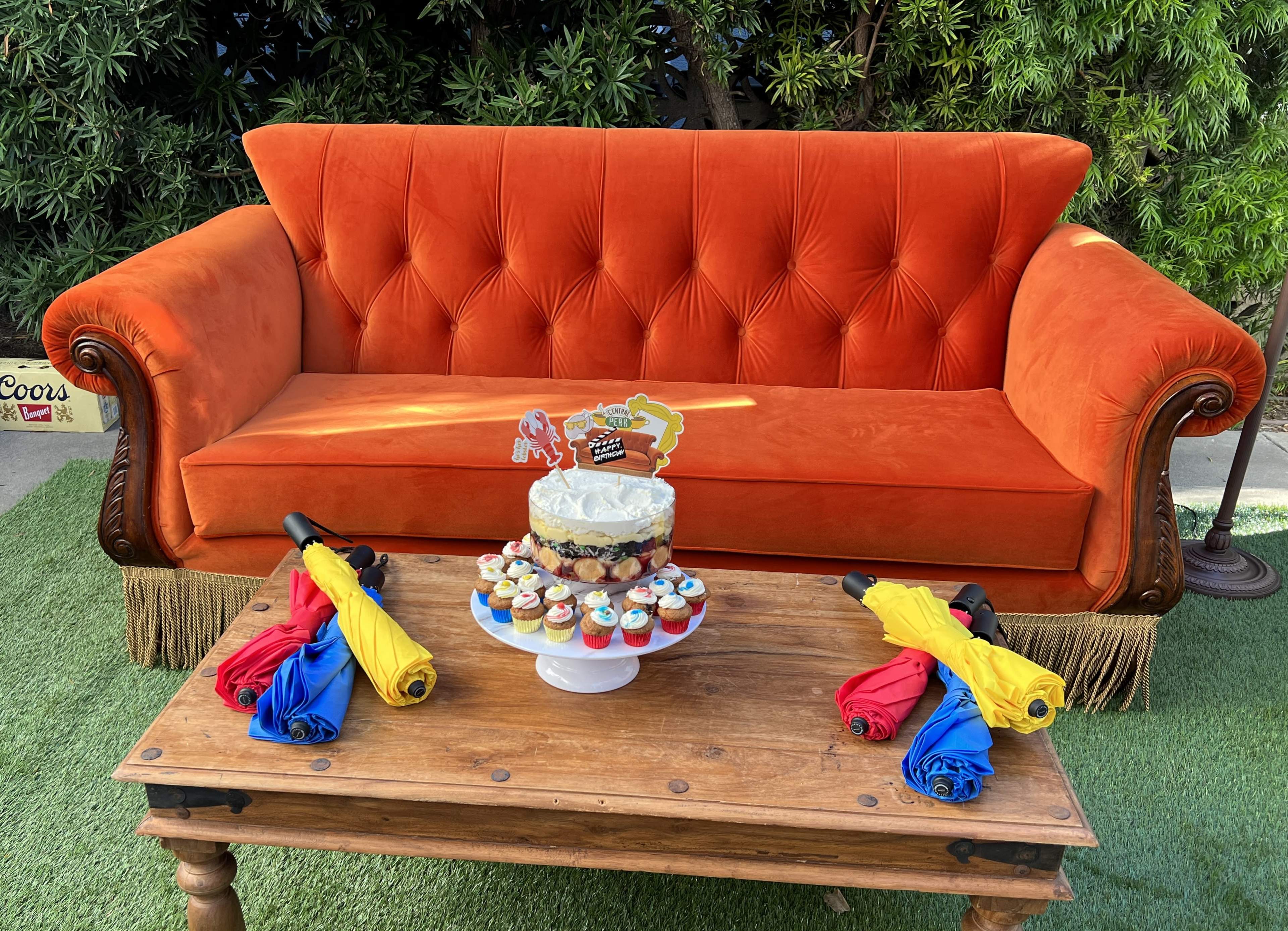 An orange velvet sofa is positioned beside a wooden table displaying a cake and colorful cupcakes, with umbrellas in red, blue, and yellow arranged nearby.