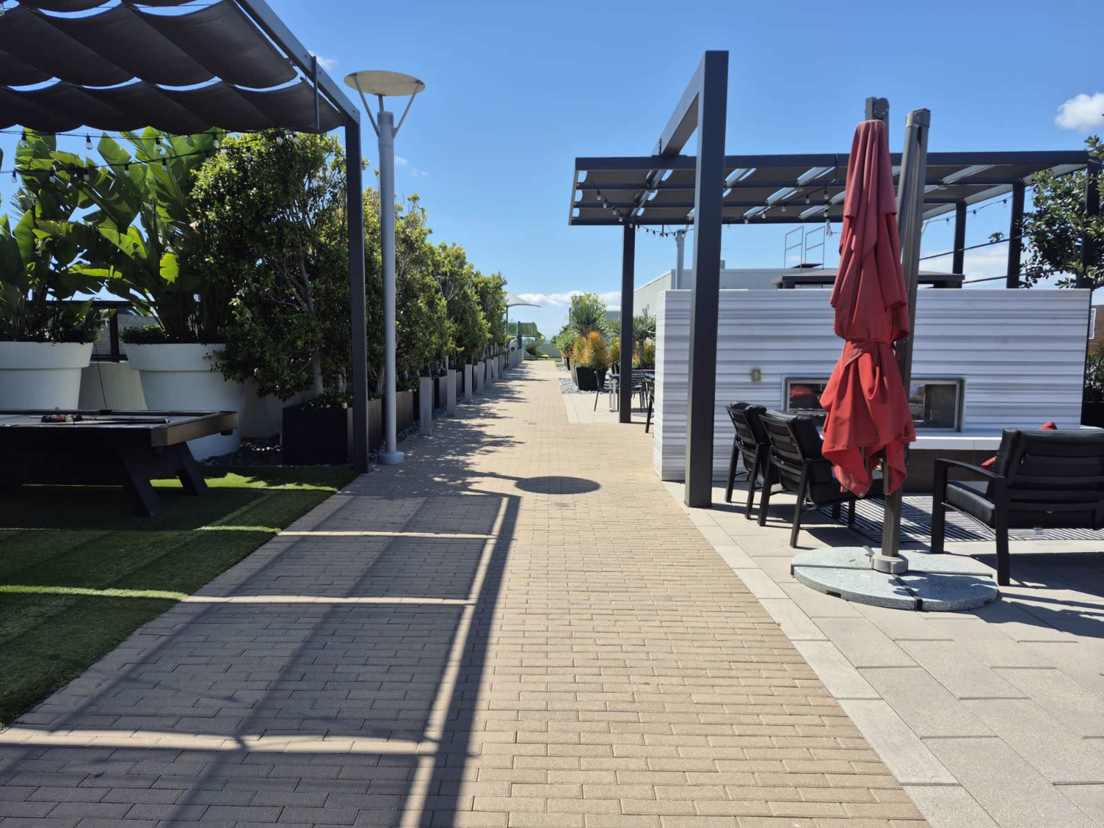 The image shows a paved walkway bordered by greenery and furniture in a modern outdoor space.