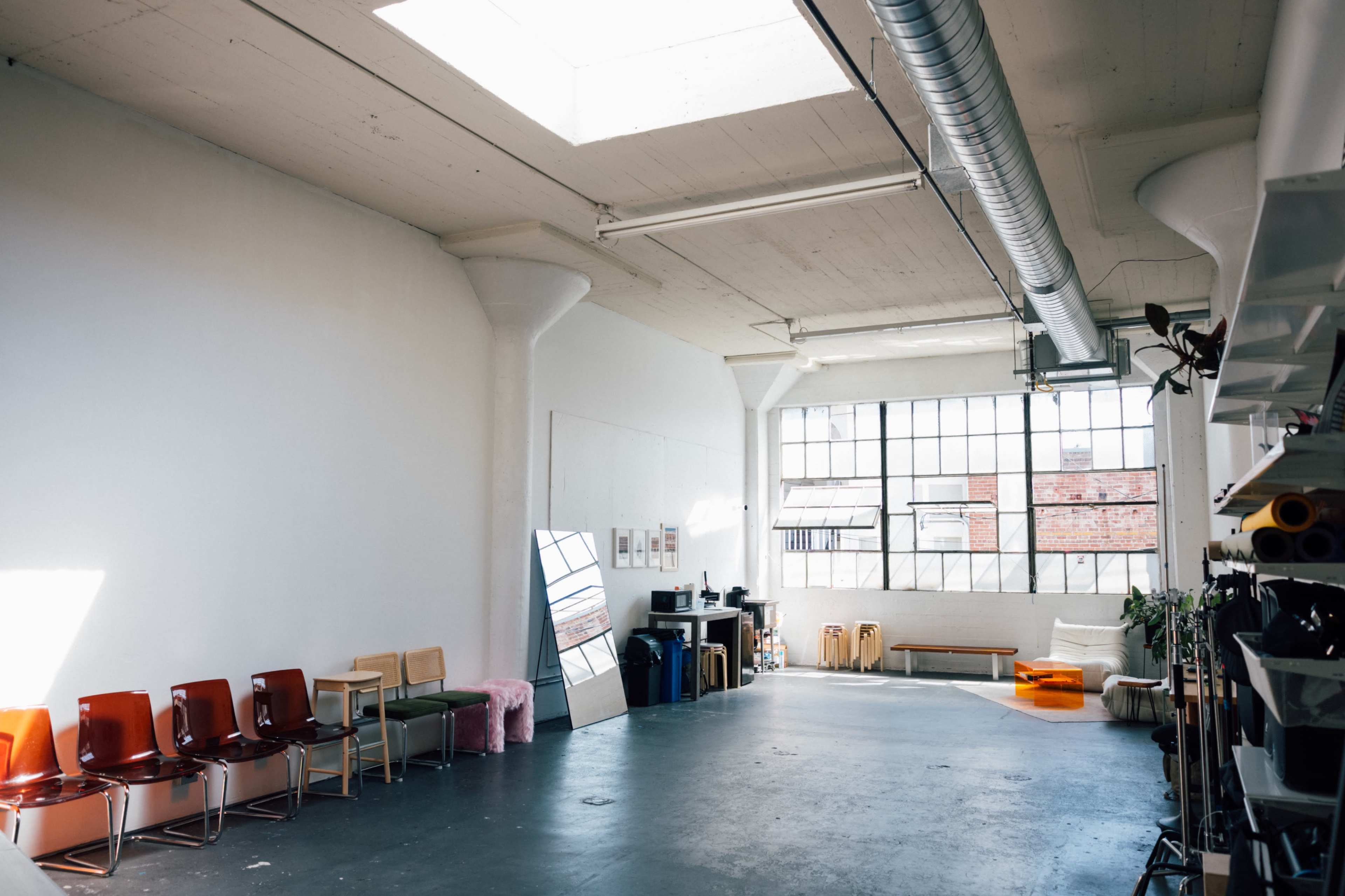 The image shows a spacious, well-lit loft featuring a row of chairs on one side and large windows providing natural light.