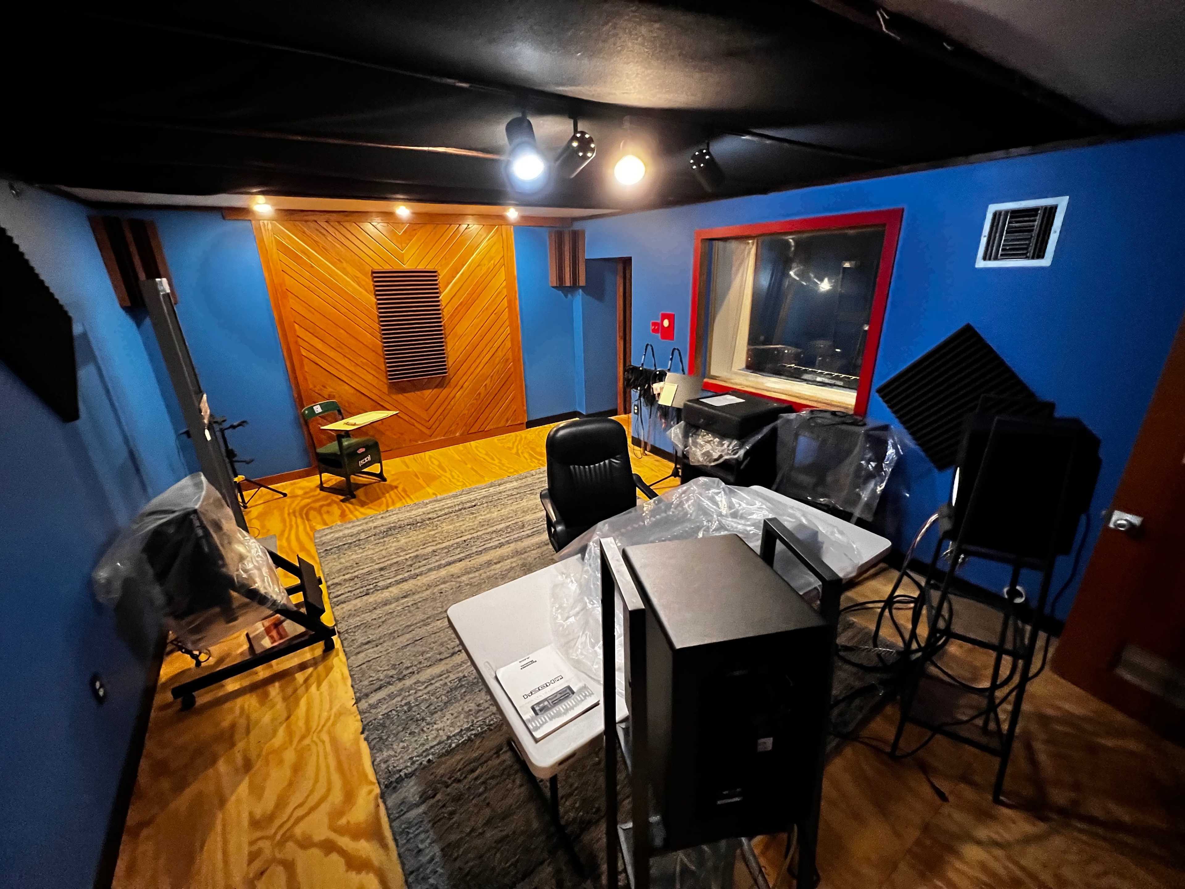 The image shows a partially furnished recording studio with blue walls, a table, chairs, and studio monitors arranged around an area intended for audio production.
