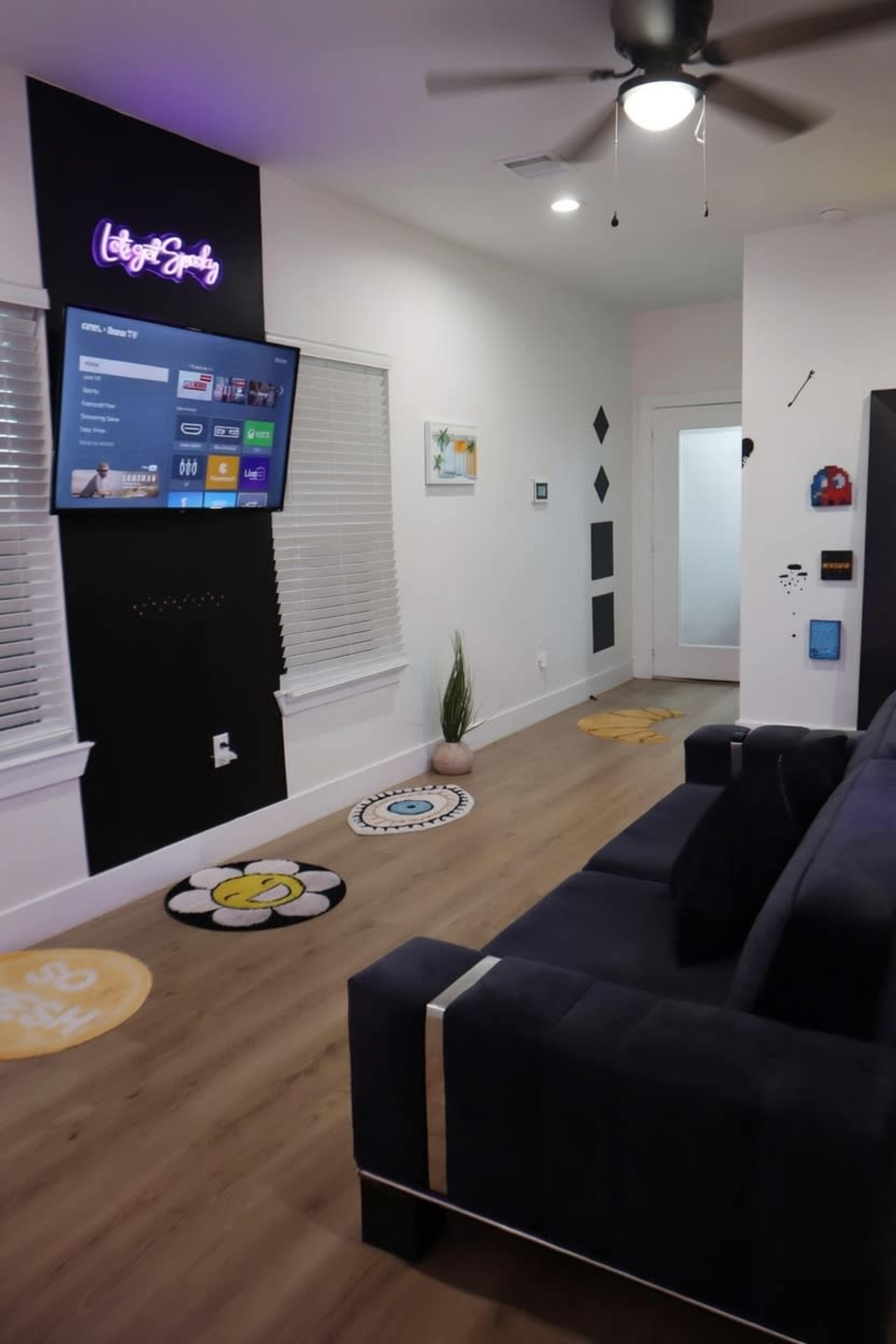 The image shows a modern living room with a dark sofa facing a wall mounted TV, decorative wall art, and colorful circular rugs on a wooden floor.